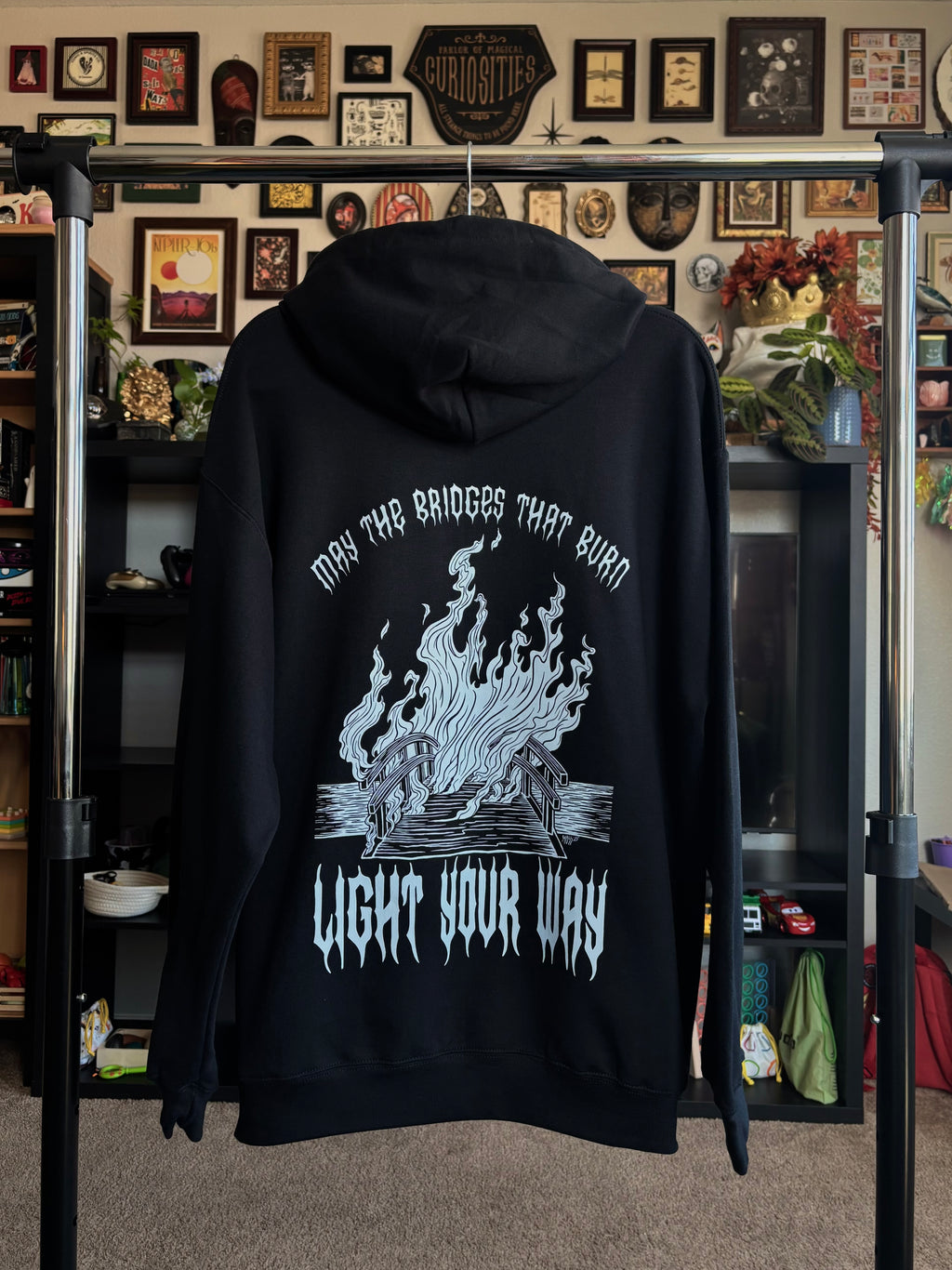 black hoodie hanging on a rack to show the full pack print of a burning bridge, surrounded by the text "MAY THE BRIDGES THAT BURN LIGHT YOUR WAY"