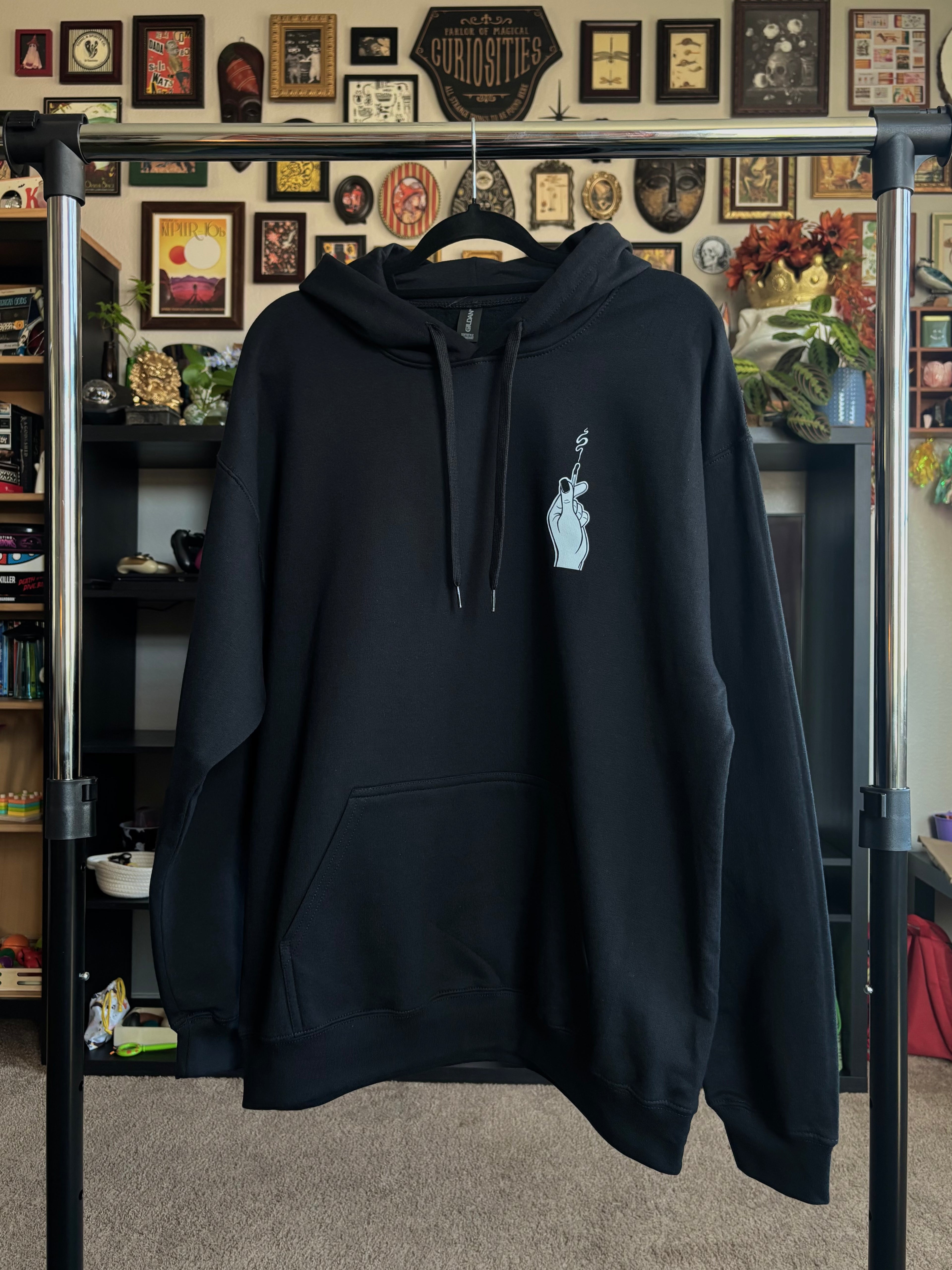 black hoodie hanging on a rack to display the front of the Burning Bridges design, which shows a hand holding a burnt match with a small spiral of smoke