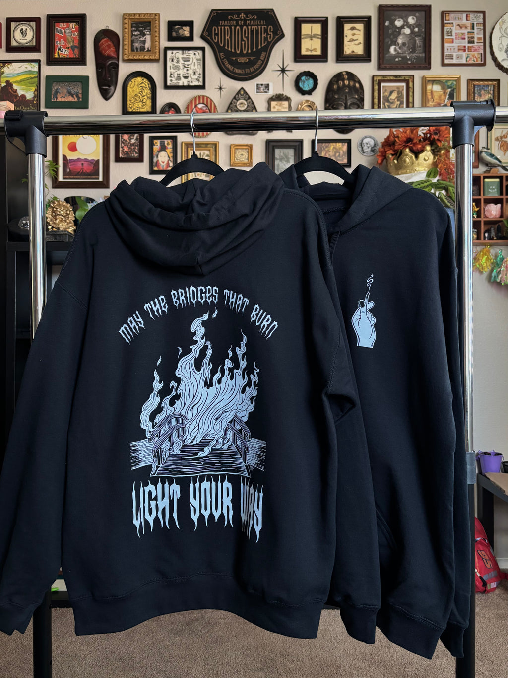 two black hoodies with the Burning Bridges print hanging on a rack to show the full back print of the burning bridge, and the club patch of the hand holding the match