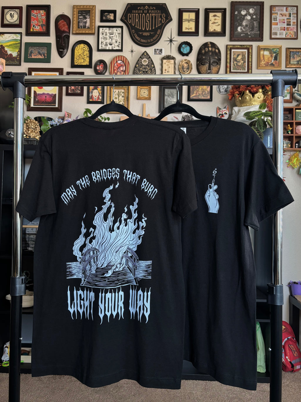 two black tees with the Burning Bridges print hanging on a rack to show the full back print of the burning bridge, and the club patch of the hand holding the match