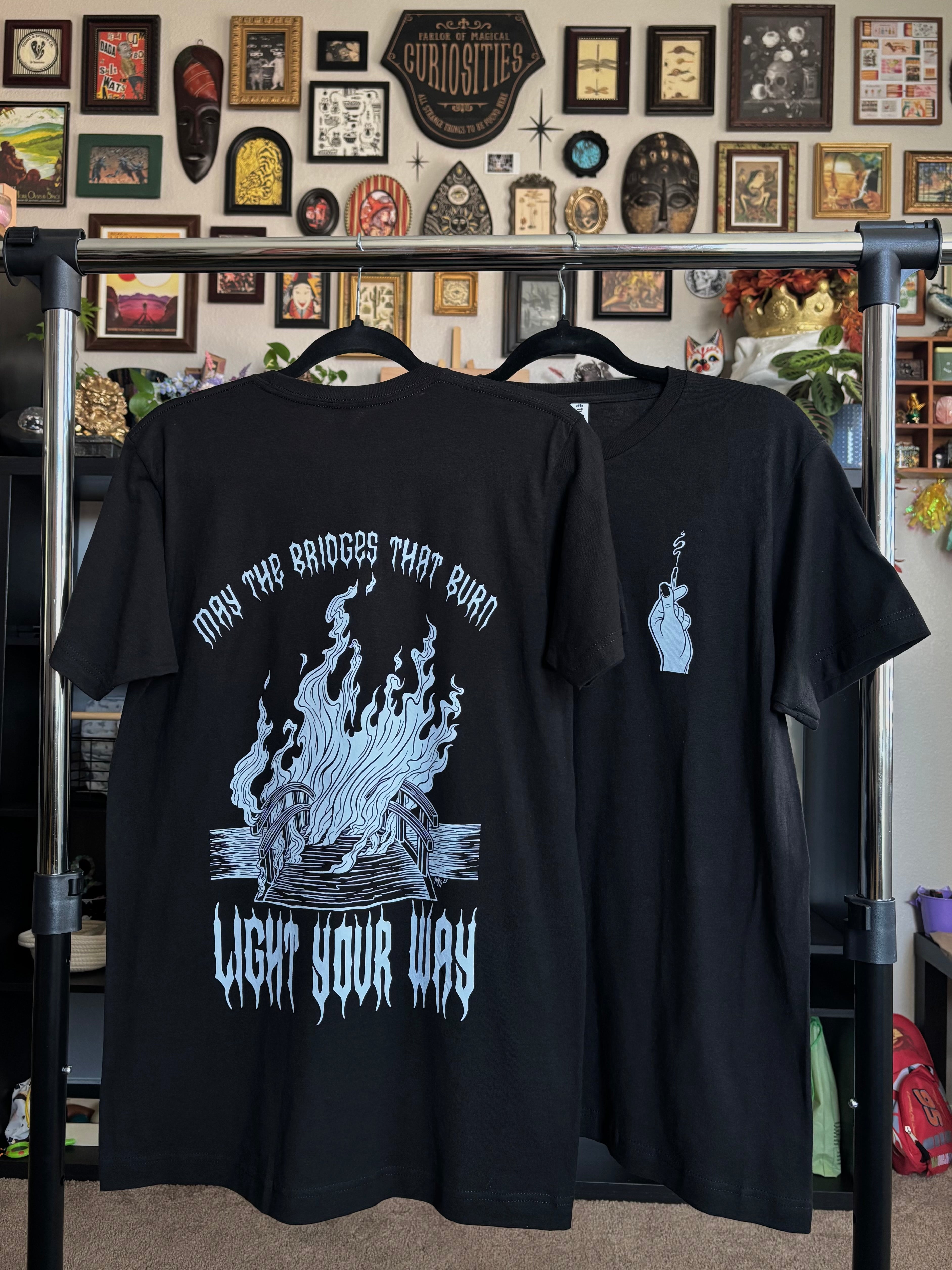 two black tees with the Burning Bridges print hanging on a rack to show the full back print of the burning bridge, and the club patch of the hand holding the match