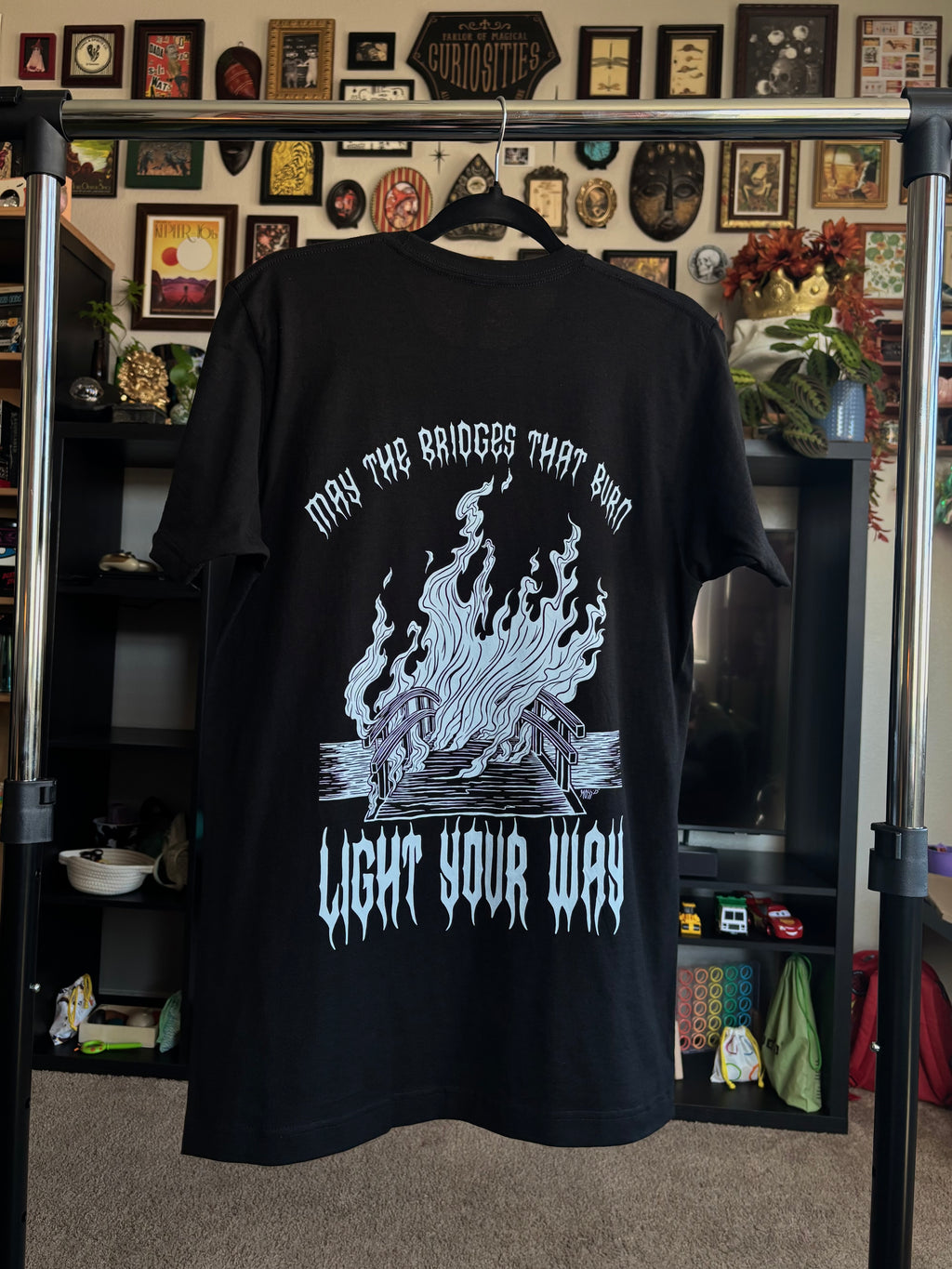 black tee hanging to display the full back print of a bridge engulfed in flames, with the word "MAY THE BRIDGES THAT BURN LIGHT YOUR WAY" printed around it