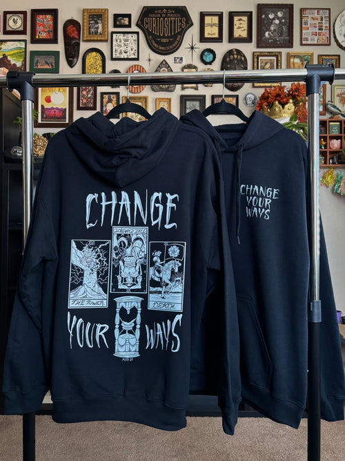 two black hoodies with the Change Your Ways tarot inspired design shown hanging on a rack, displaying the full-back print and the club patch on the front