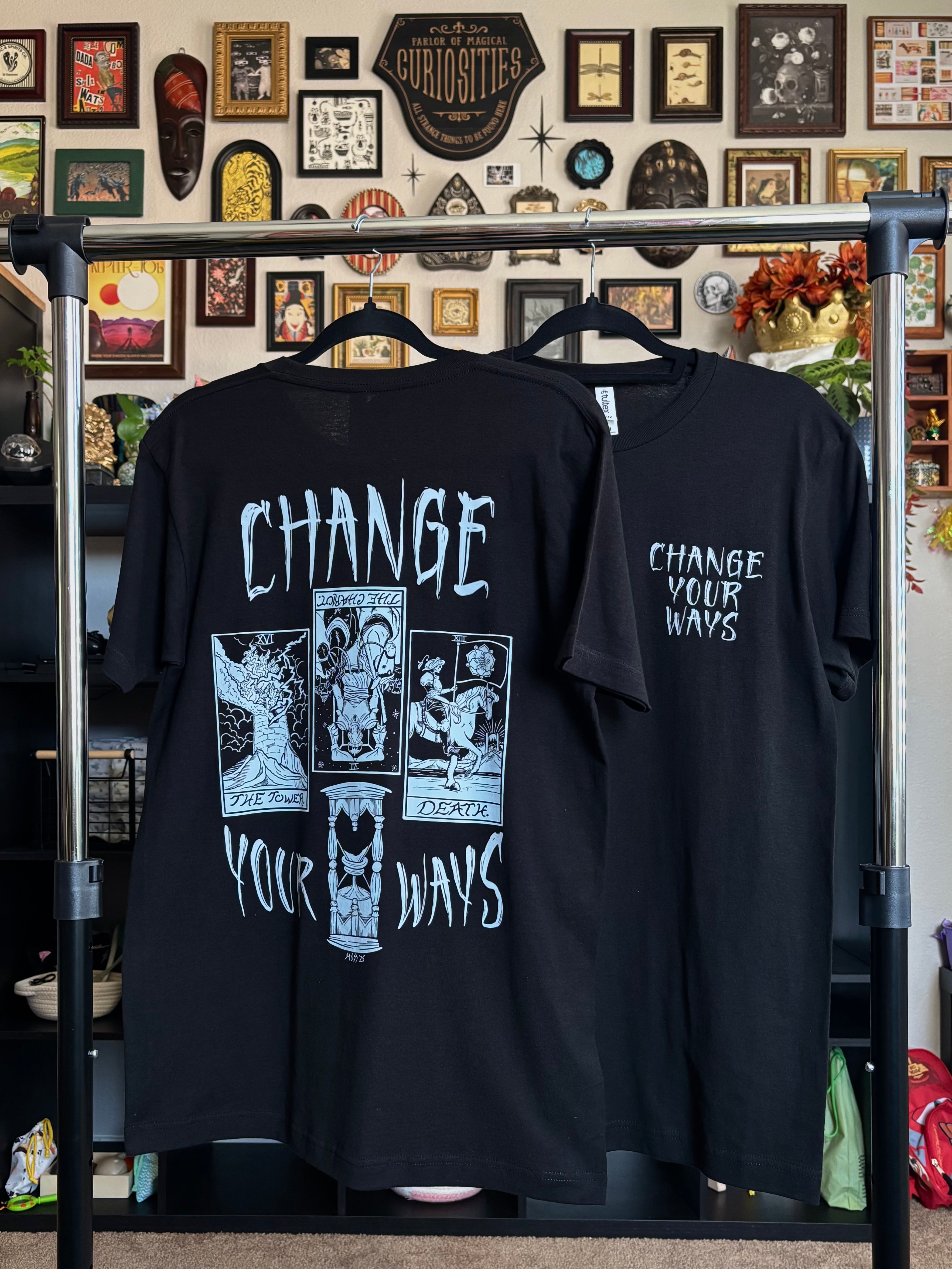 two black tees with the Change Your Ways tarot inspired design shown hanging on a rack, displaying the full-back print and the club patch on the front
