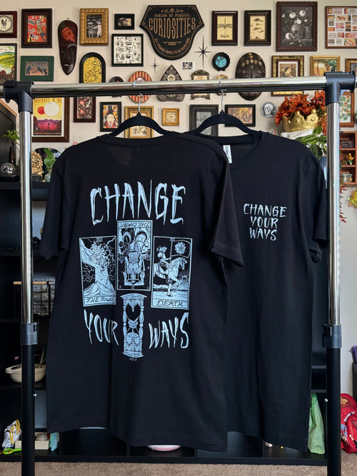 two black tees with the Change Your Ways tarot inspired design shown hanging on a rack, displaying the full-back print and the club patch on the front