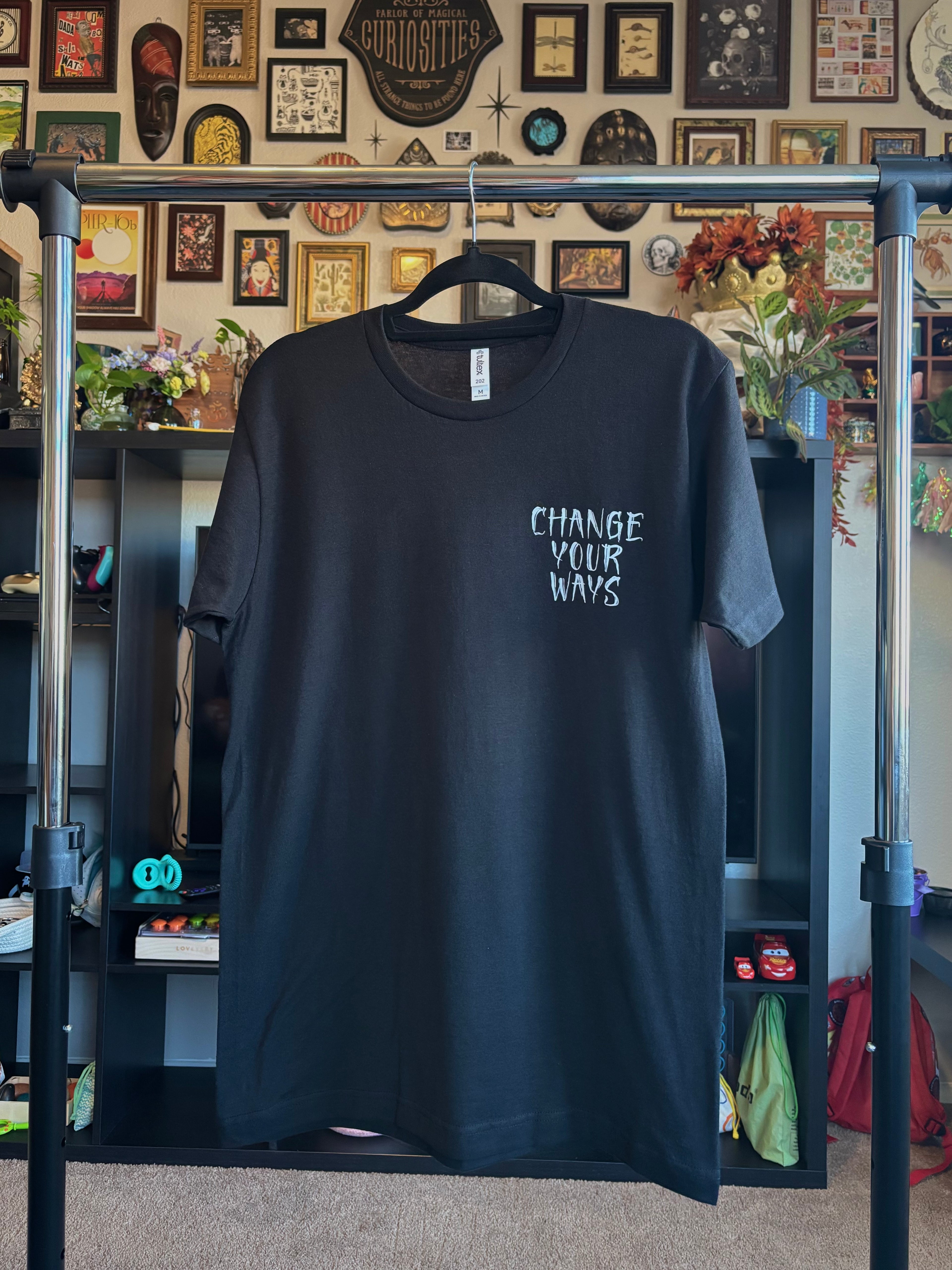 the Change Your Ways tarot inspired design printed on a black t-shirt, hanging on a rack to show the front with a club patch print