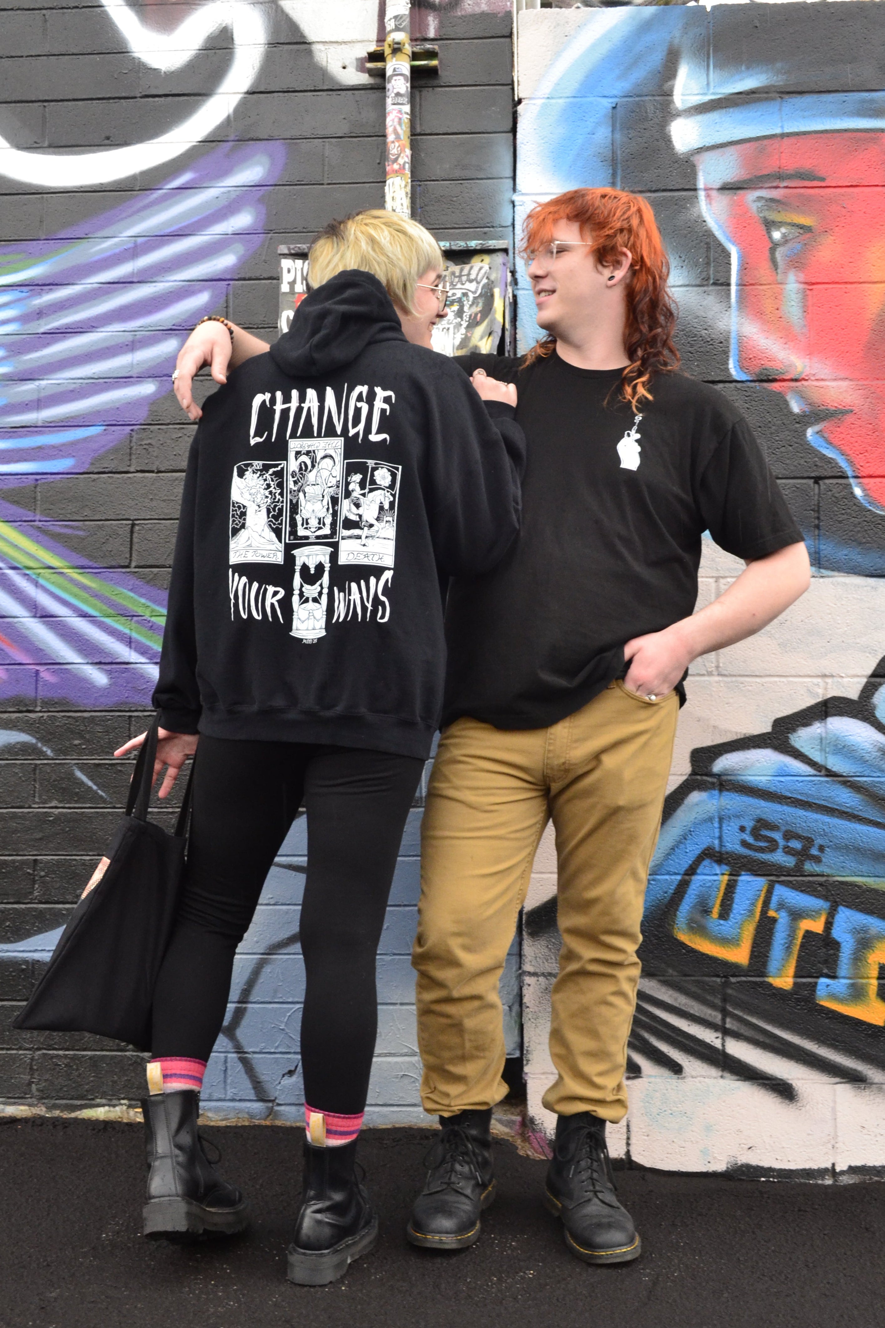 two models stand, one with their back to the camera to show off the full print on the Change Your Ways hoodie while the other stands facing the camera to show the club patch on the Burning Bridges t-shirt