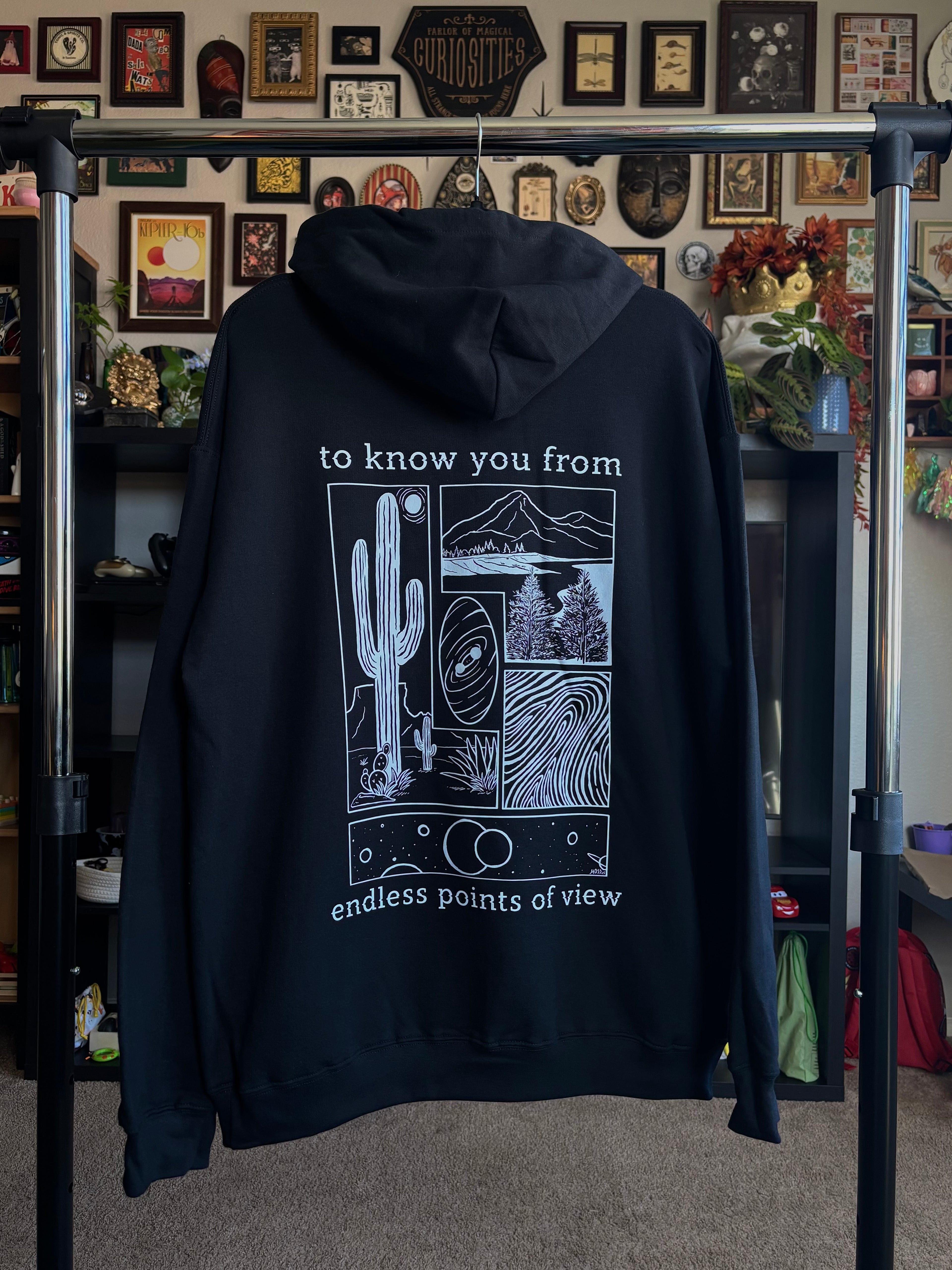 black hoodie hanging on a rack to display the full print on the back, including a desert scene, a river flowing through a valley, a closeup of a finger print, and images of the cosmos. "to know you from endless points of view" is printed around the scenes
