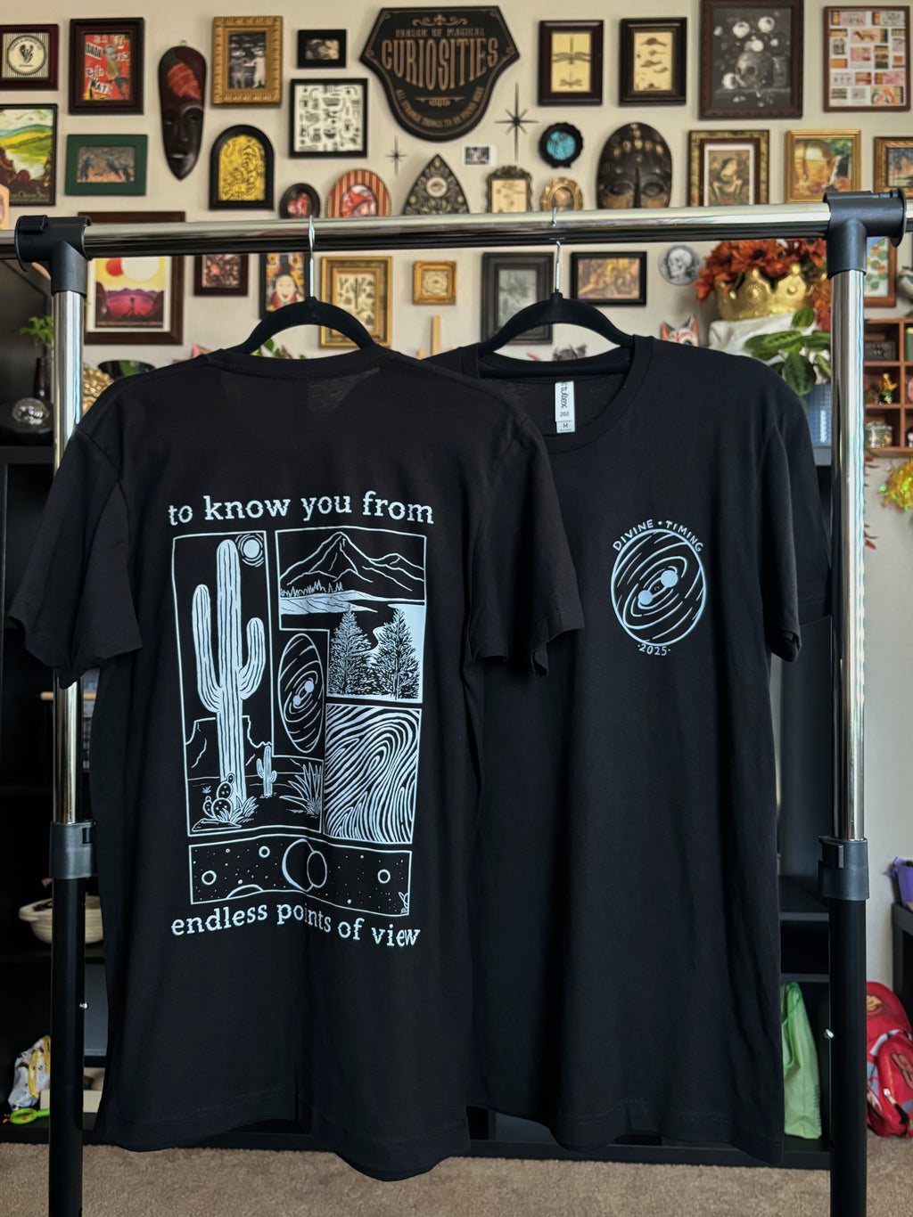 two black tees hanging on a rack to show the full back print and the front club patch for the Endless Points of View design