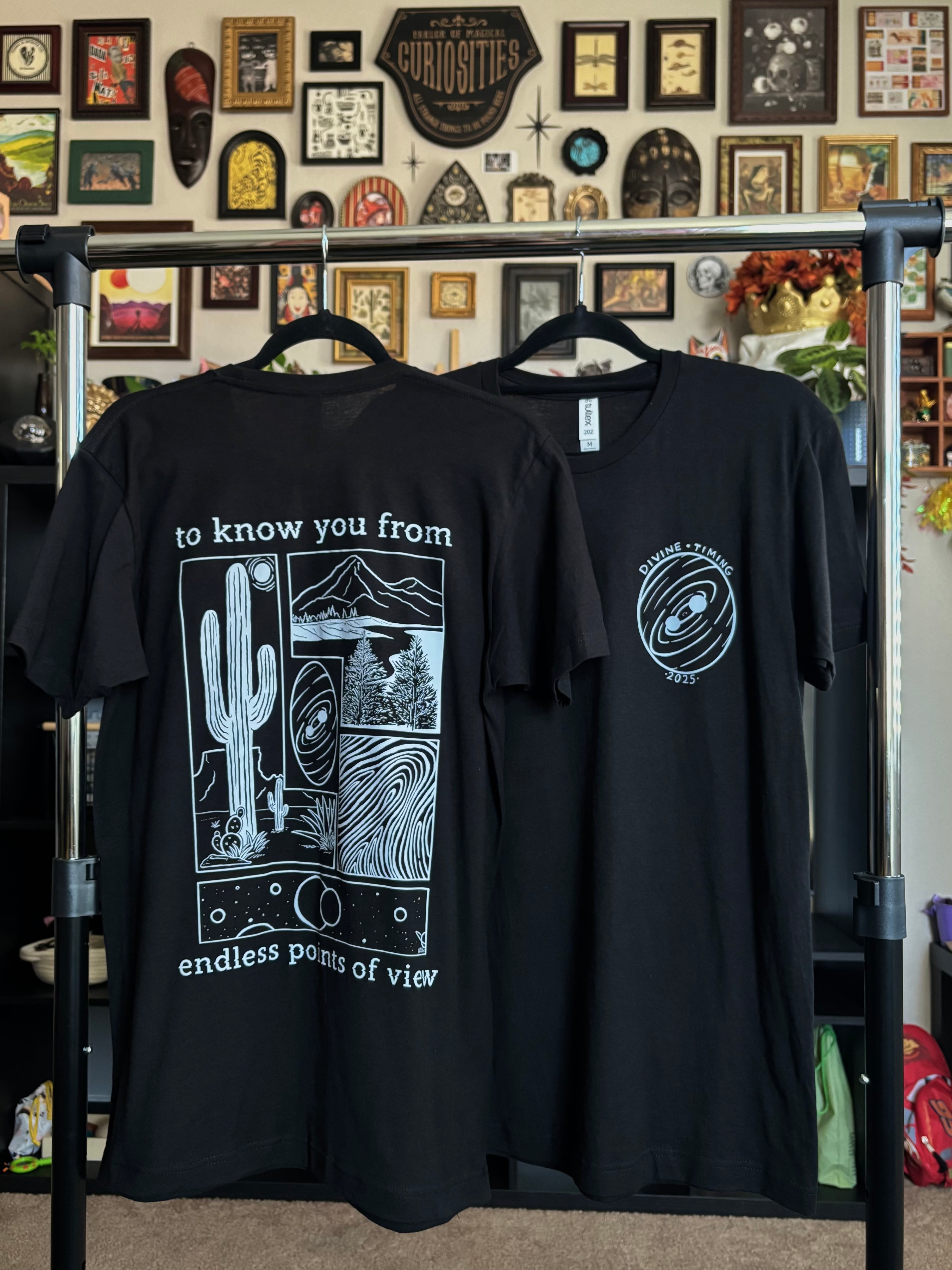 two black tees hanging on a rack to show the full back print and the front club patch for the Endless Points of View design