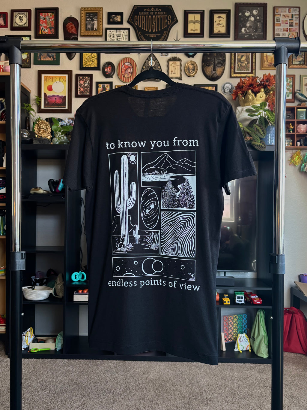 black tee hanging on a rack to display the full print on the back, including a desert scene, a river flowing through a valley, a closeup of a finger print, and images of the cosmos. "to know you from endless points of view" is printed around the scenes