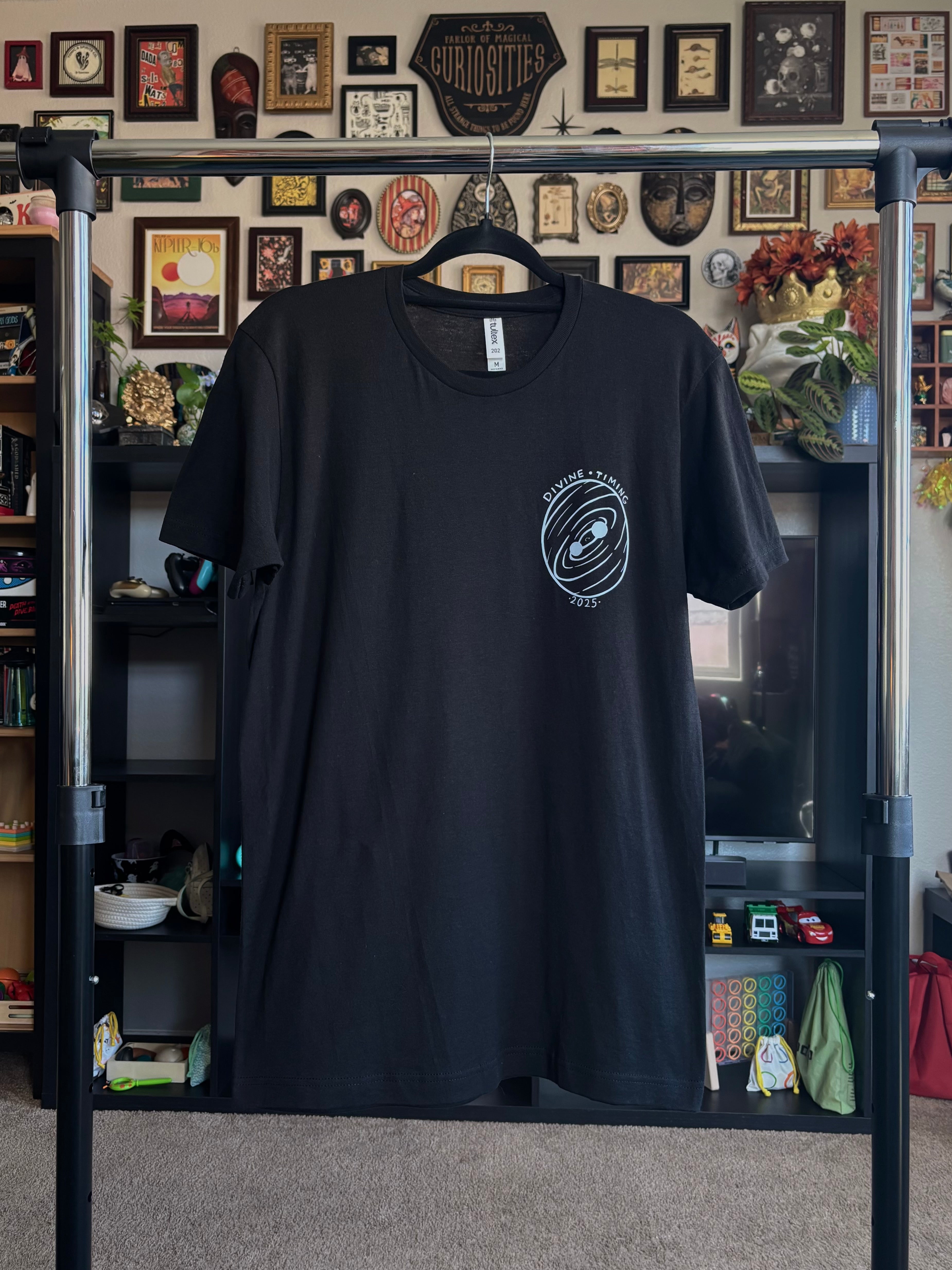 black tee hanging on a rack to show the front with the Divine Timing Binary Star badge printed as a club patch