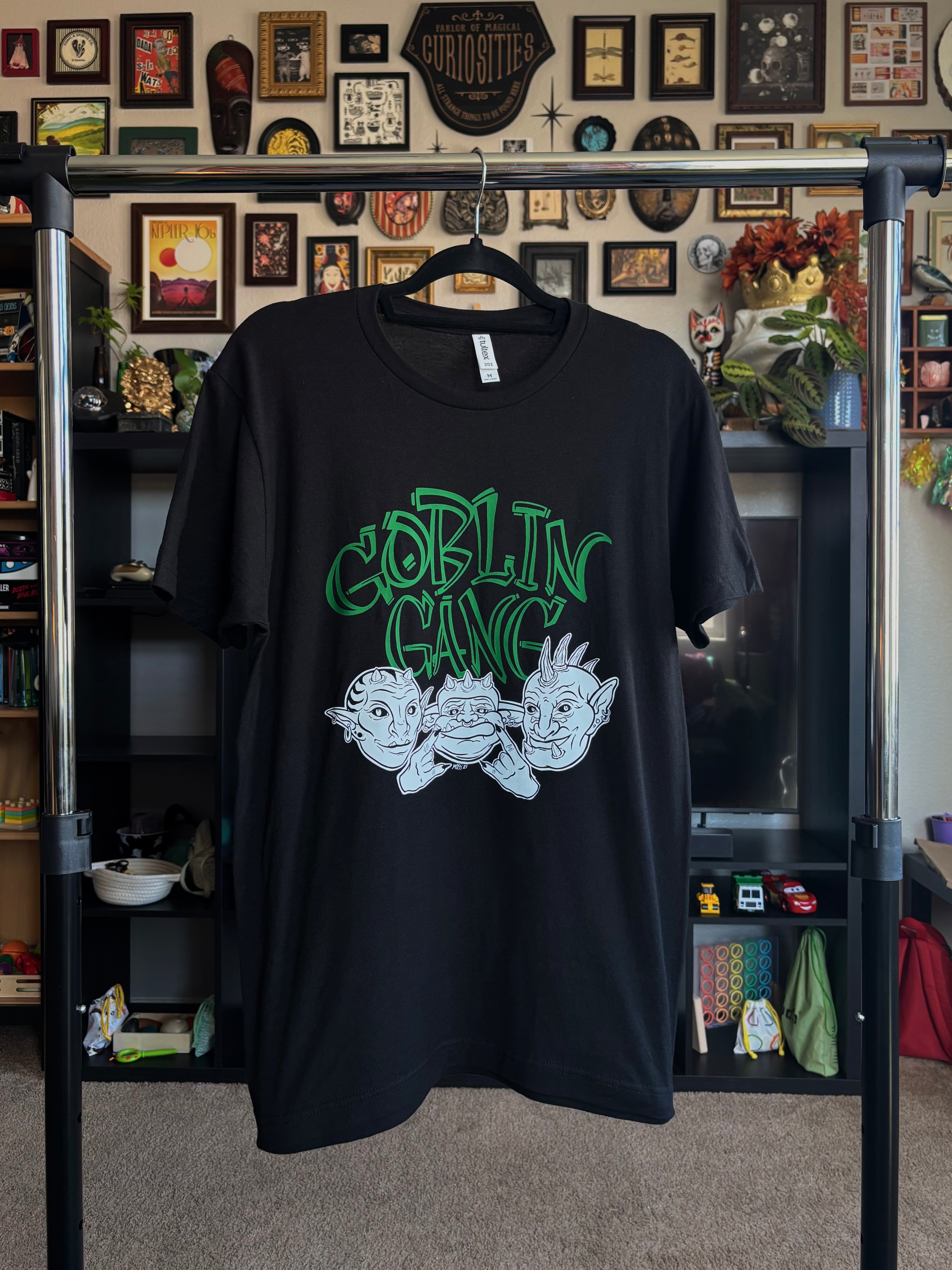 black tee with "GOBLIN GANG" printed in green across the chest, and the faces of three goblins printed in black and white below, hanging on a rack