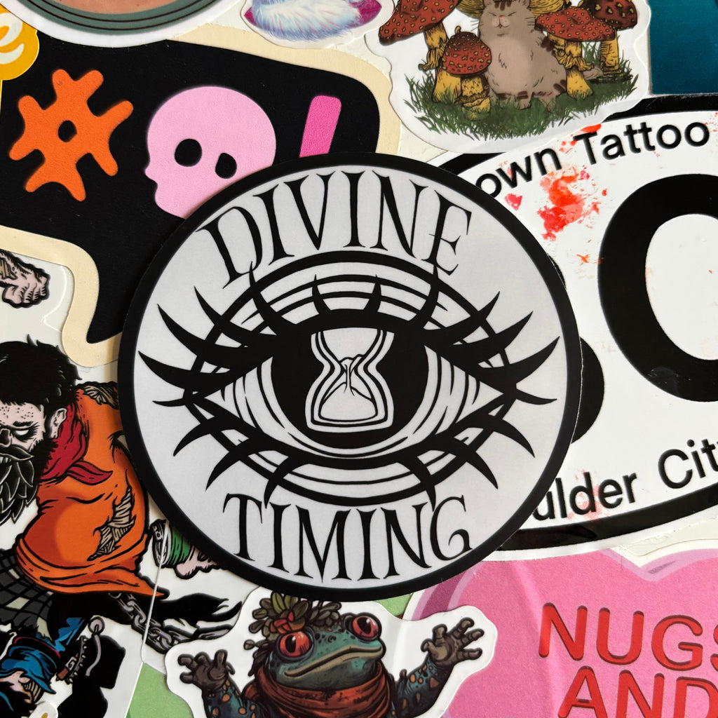 sticker of the Divine Timing logo which shows an hourglass in an eye, with the sand almost about to run out