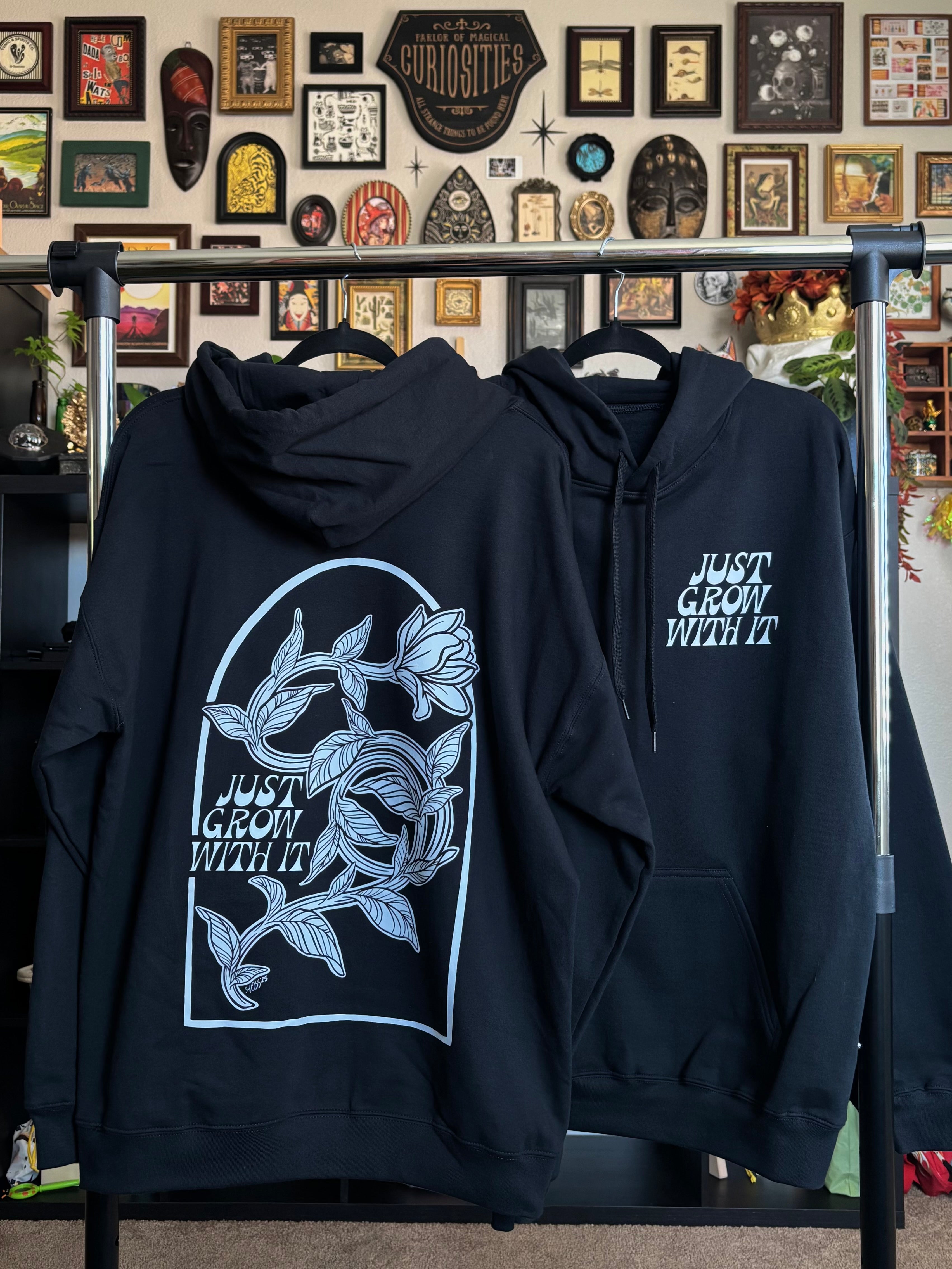 two black hoodies hanging on a rack to show the full back print of the twisting flower and the "JUST GROW WITH IT" club patch on the front