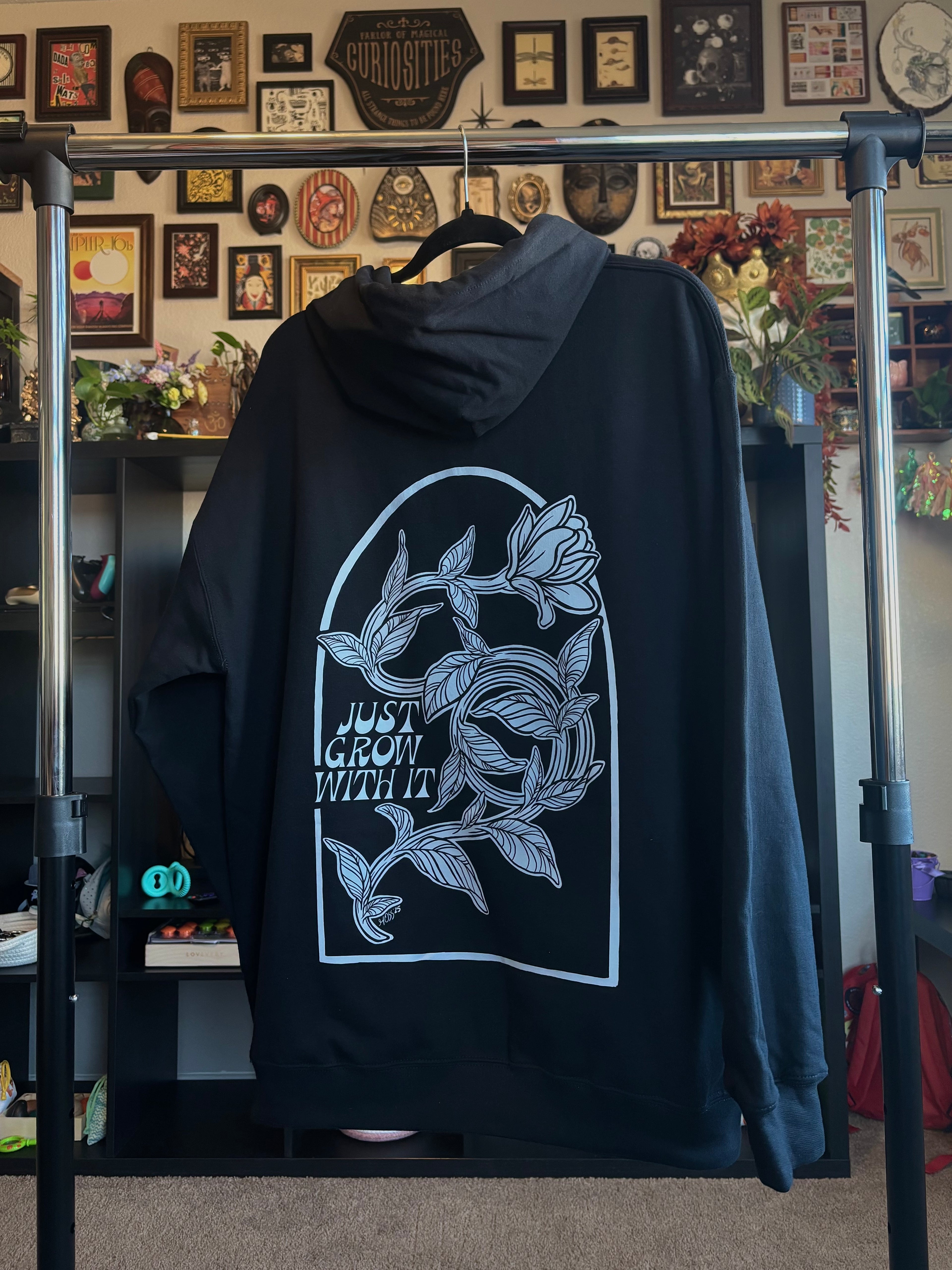 black hoodie hanging on a rack to show off the full back print of a defiant flower twisting and turning as it grows