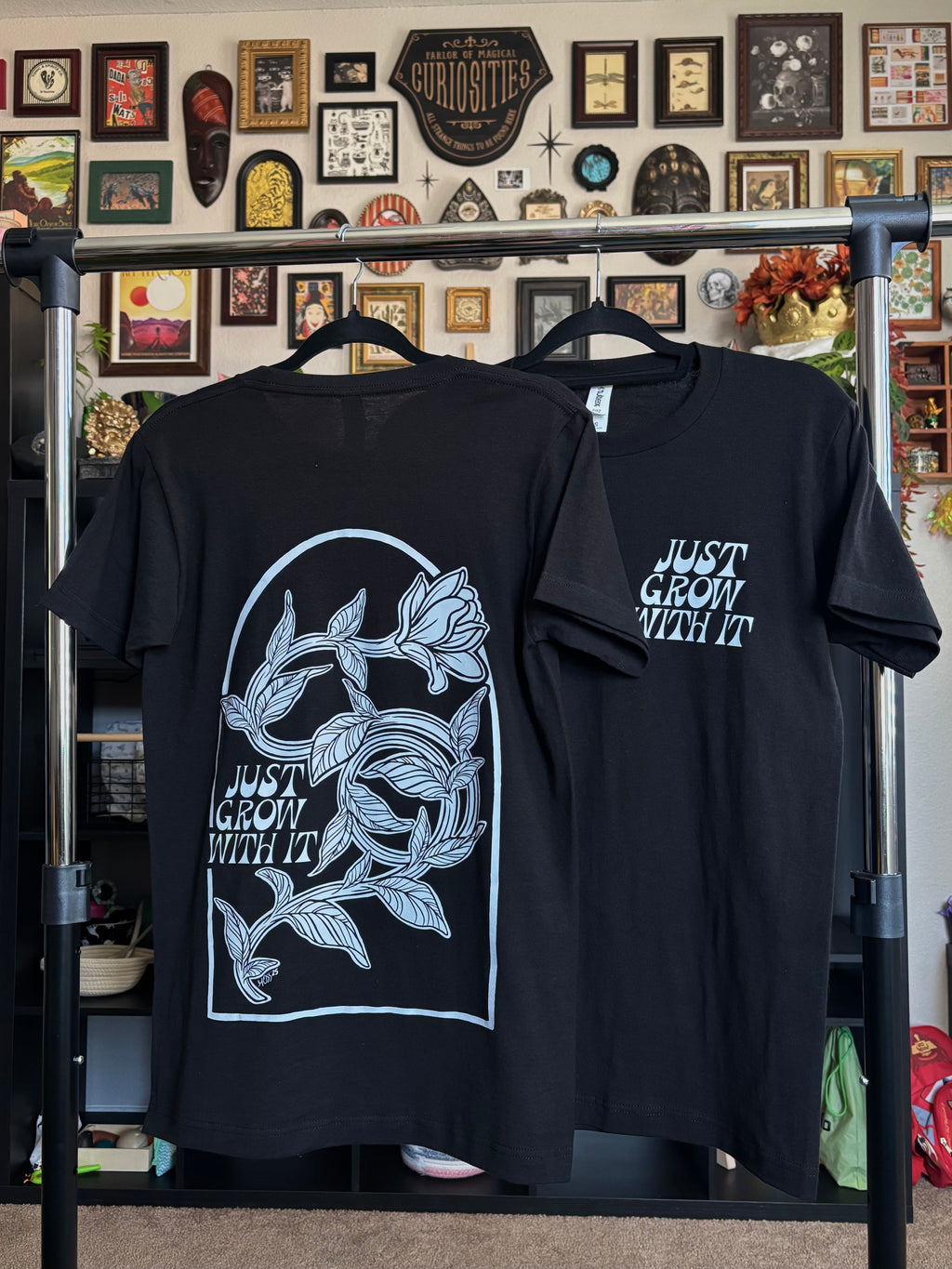 two black tees hanging on a rack to show the full back print of the twisting flower and the "JUST GROW WITH IT" club patch on the front