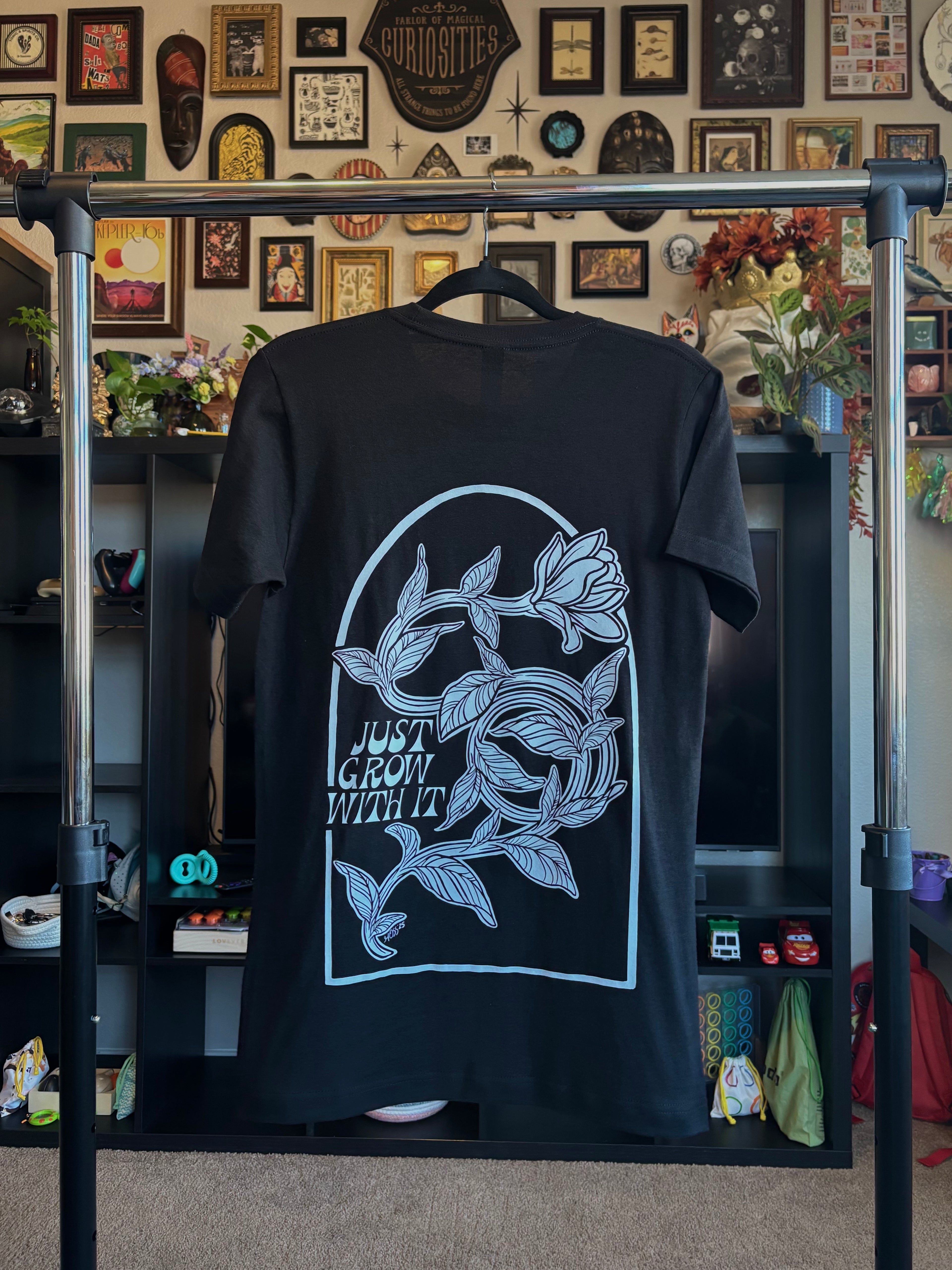 black t-shirt hanging on a rack to show off the full back print of a defiant flower twisting and turning as it grows