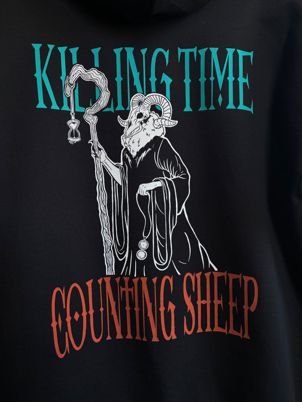 closeup image of the Killing Time design printed across the back of a black hoodie, showing the Keeper of Time surrounded by the text "Killing Time, Counting Sheep"