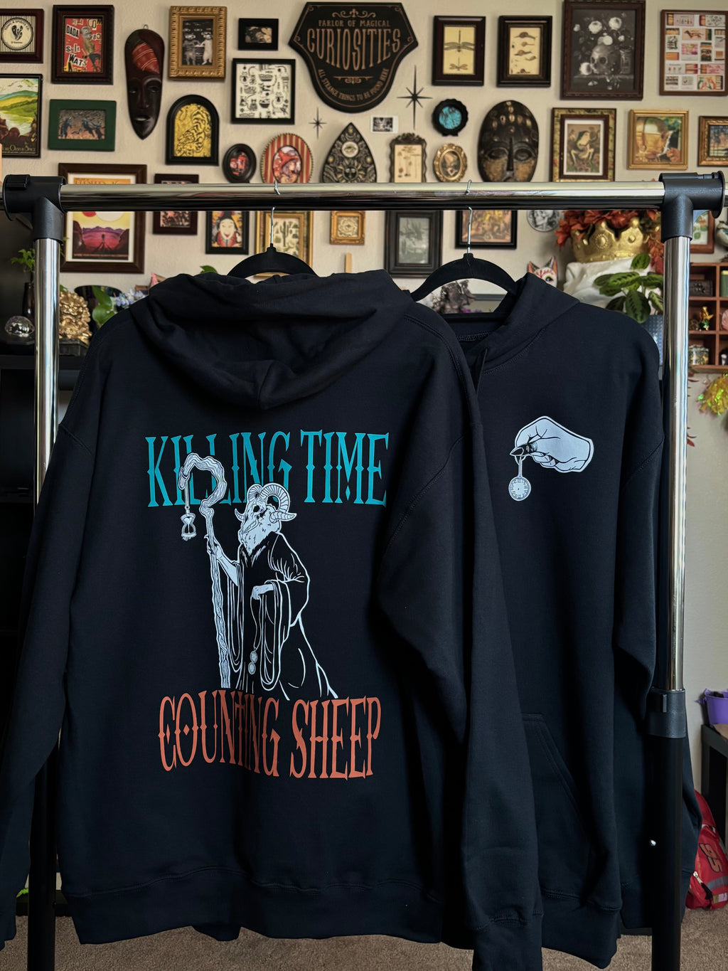 two black hoodies hanging on a rack to show the front and back of the Killing Time design, with the Keeper of Time printed on the back surrounded by the words "KILLING TIME, COUNTING SHEEP" and a hand holding a small pocket watch printed as a club patch on the front