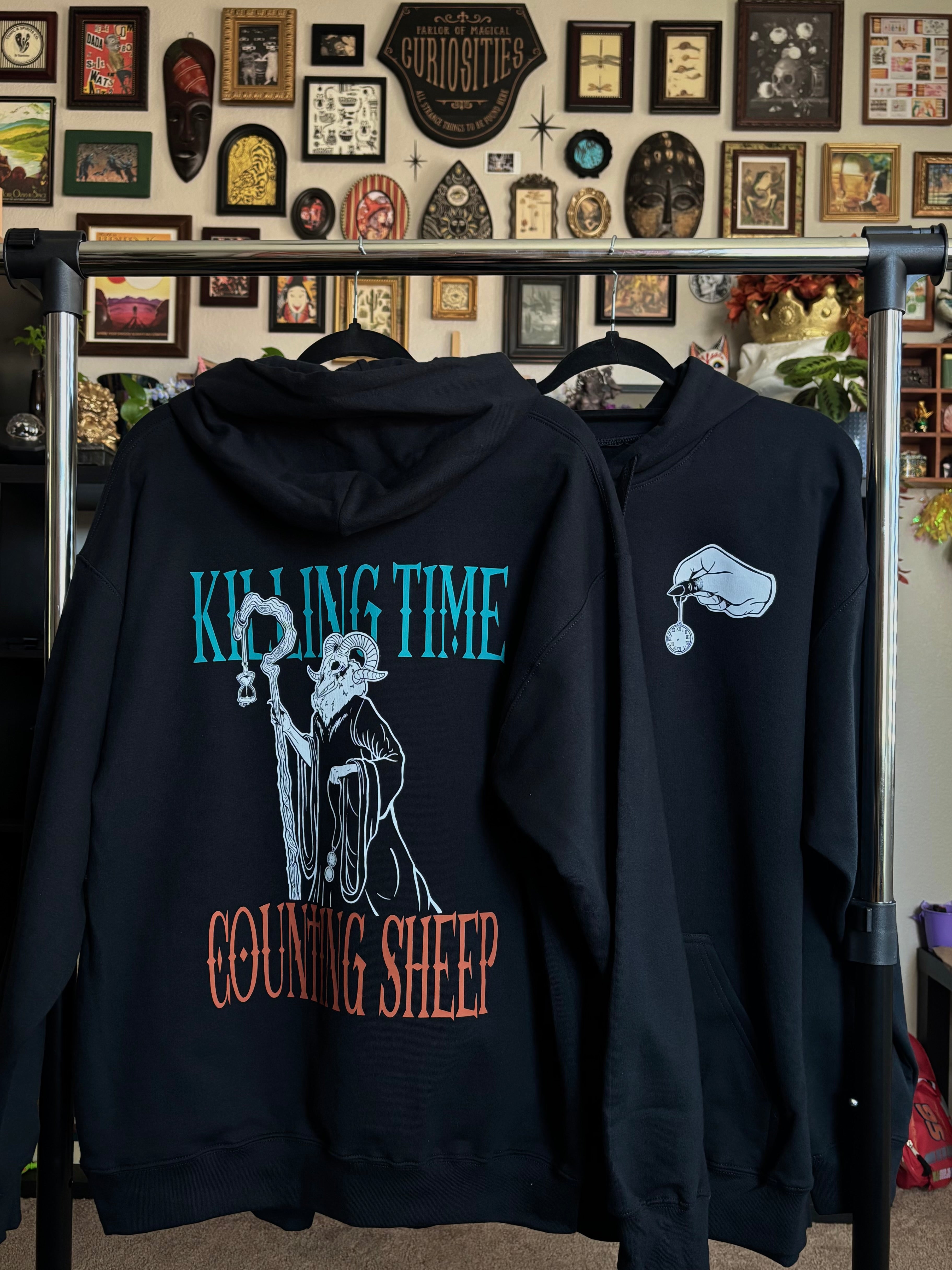 two black hoodies hanging on a rack to show the front and back of the Killing Time design, with the Keeper of Time printed on the back surrounded by the words "KILLING TIME, COUNTING SHEEP" and a hand holding a small pocket watch printed as a club patch on the front