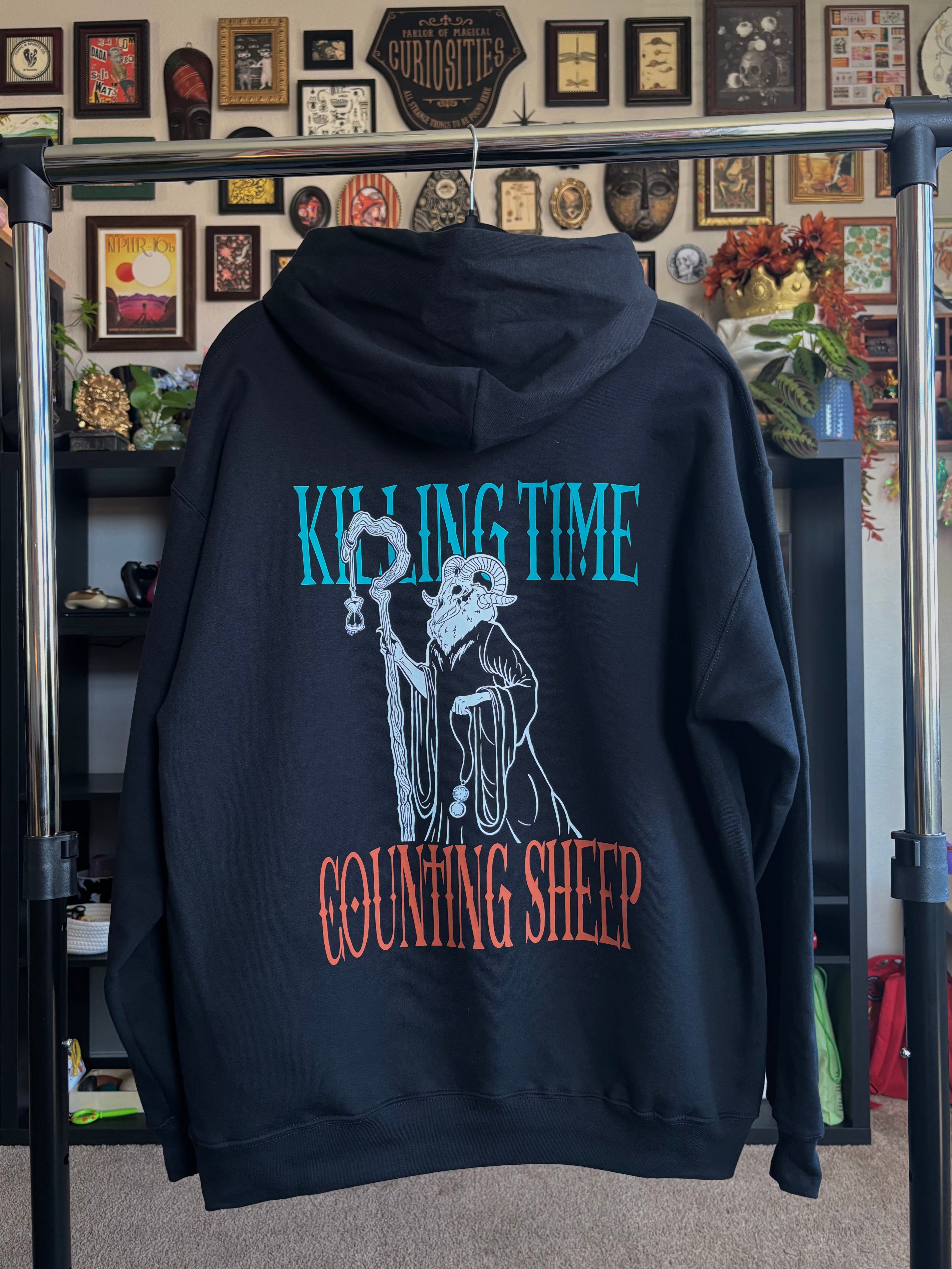 black hoodie hanging on a rack to show the full back print, with the Keeper of Time surrounded by the text 'Killing Time, Counting Sheep' 