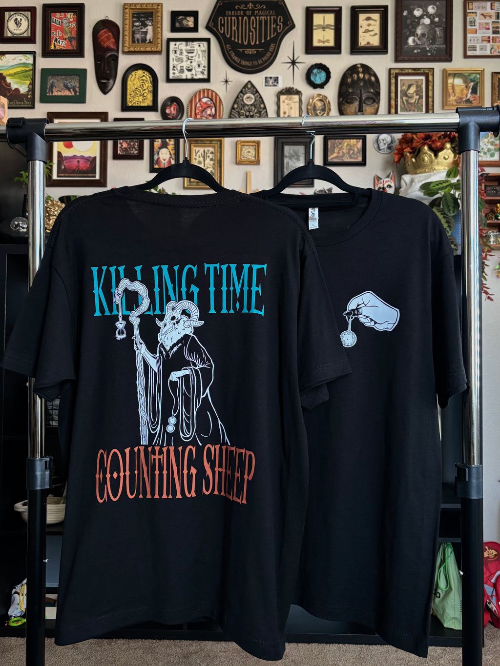 two black tees hanging on a rack to show the front and back of the Killing Time design, with the Keeper of Time printed on the back surrounded by the words "KILLING TIME, COUNTING SHEEP" and a hand holding a small pocket watch printed as a club patch on the front