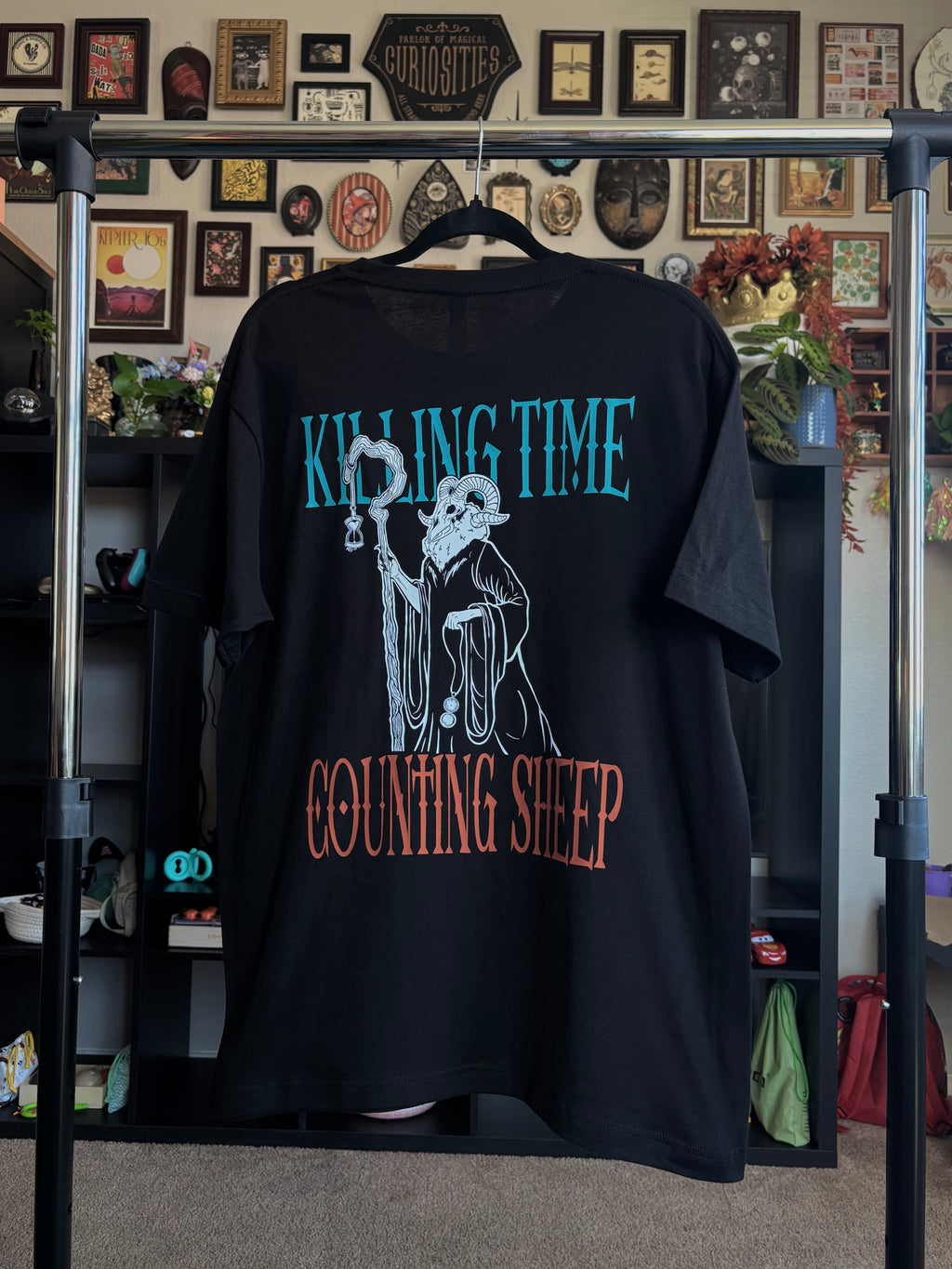 black tee hanging on a rack to show the full back print, with the Keeper of Time surrounded by the text 'Killing Time, Counting Sheep' 