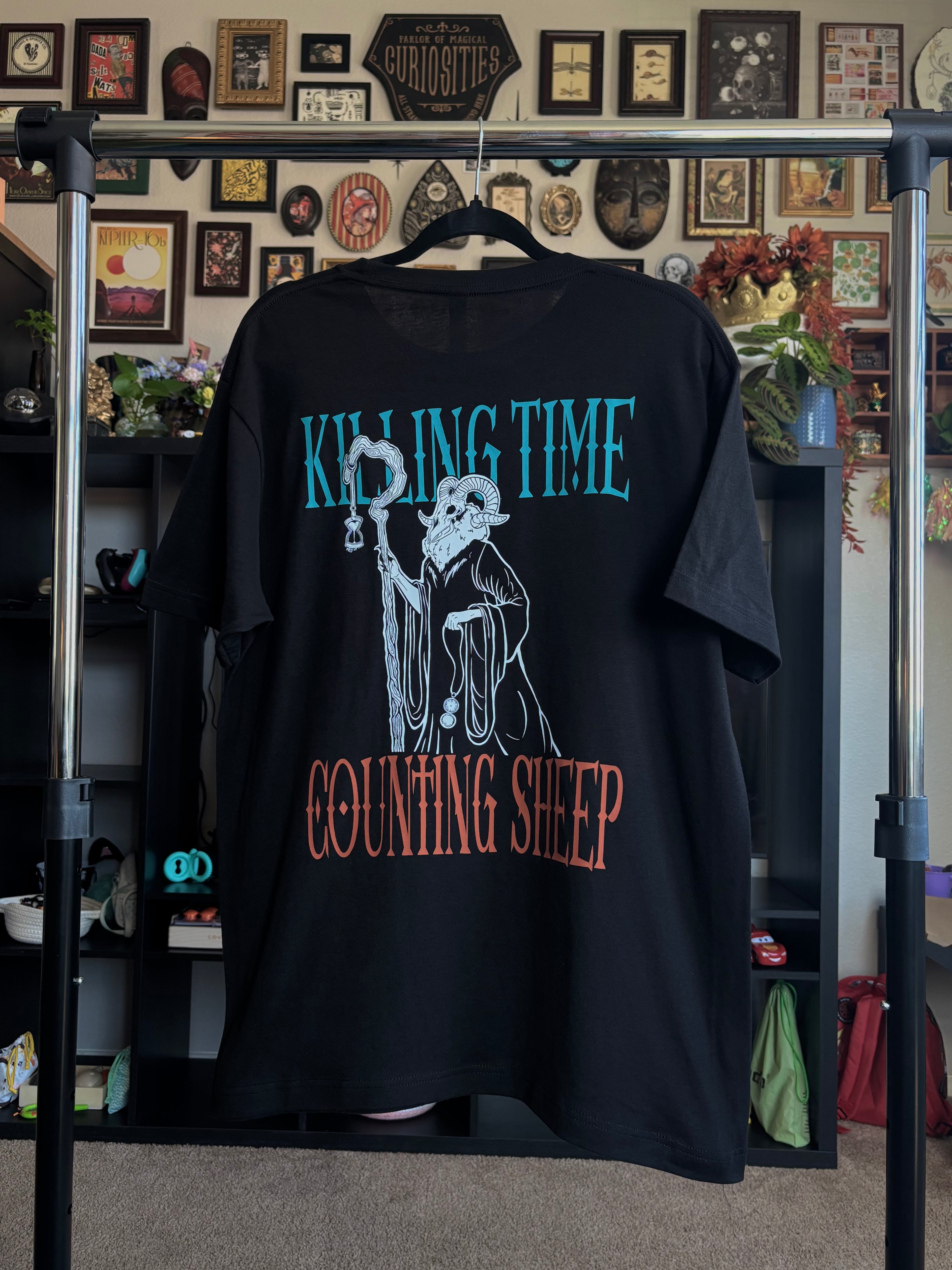 black tee hanging on a rack to show the full back print, with the Keeper of Time surrounded by the text 'Killing Time, Counting Sheep' 