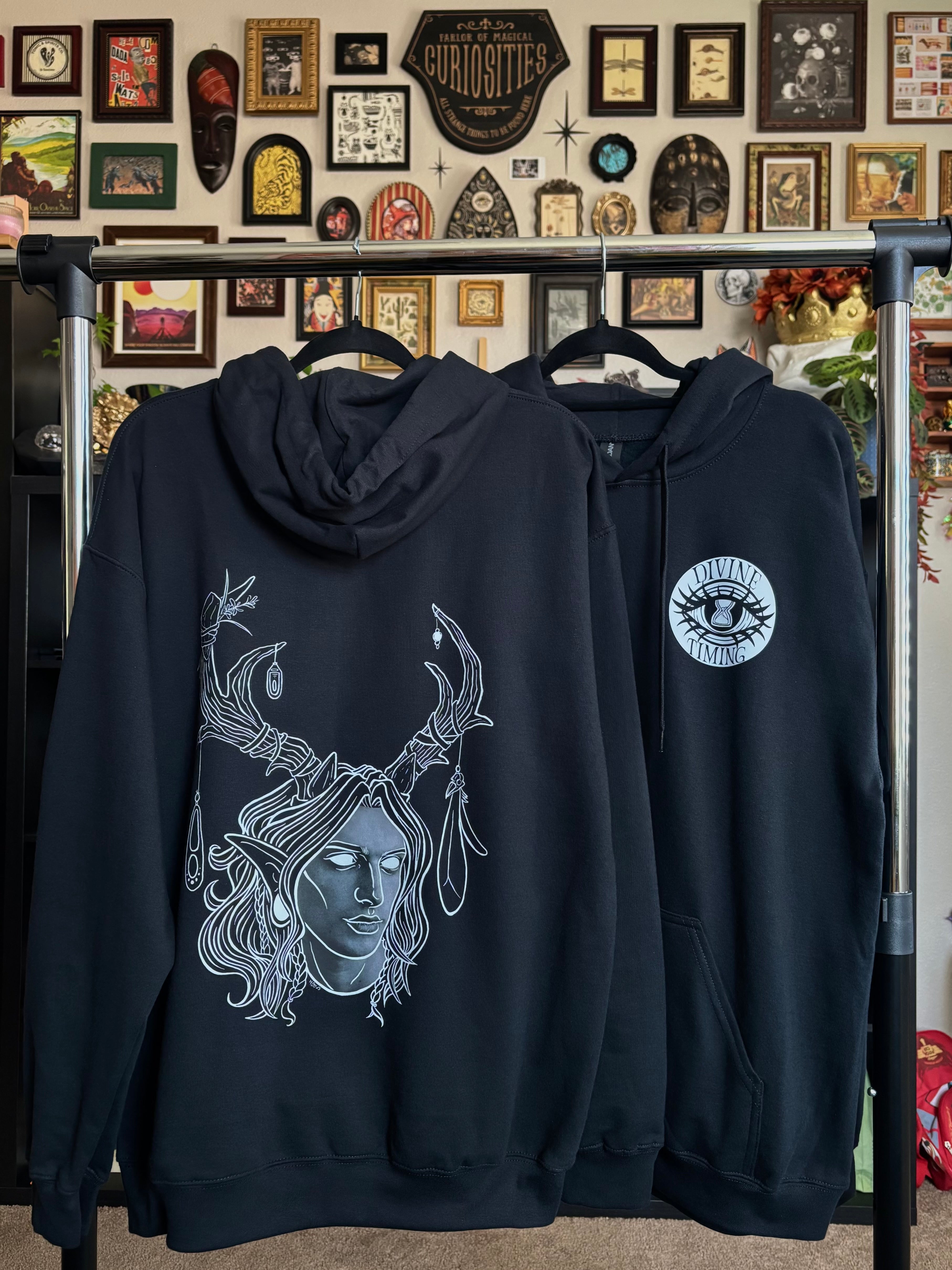 two black hoodies hanging on a rack to display the King of the Forest, a Celtic inspired fae lord, printed across the back and the Divine Timing Hourglass Eye printed as a club patch on the front