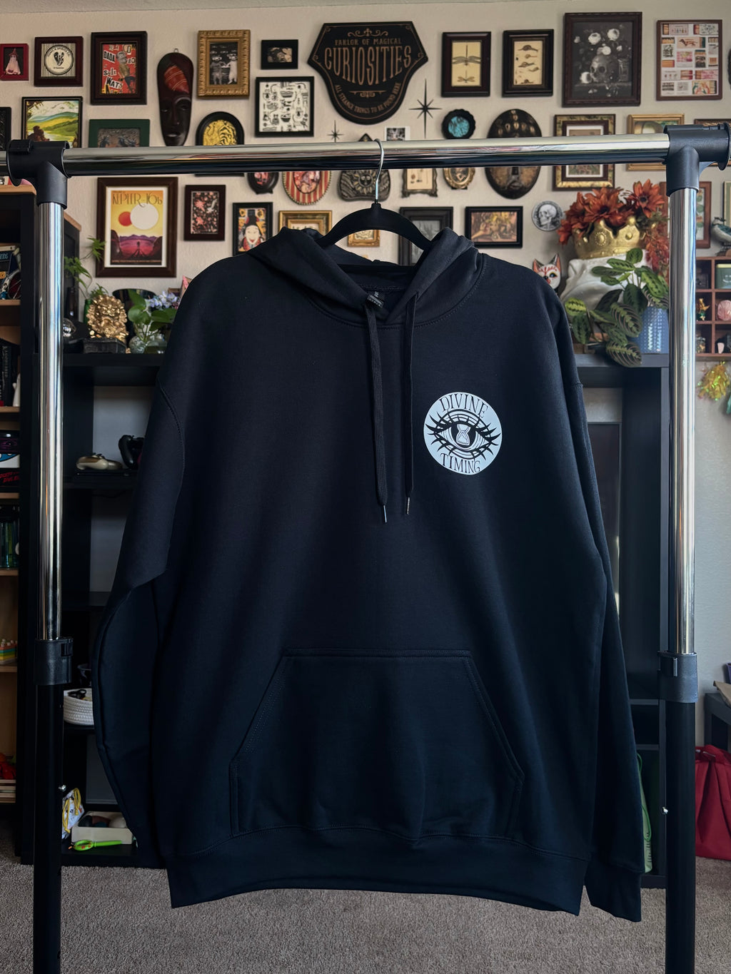 black hoodie hanging on a rack to show the front with the Divine Timing Hourglass Eye logo printed as a club patch