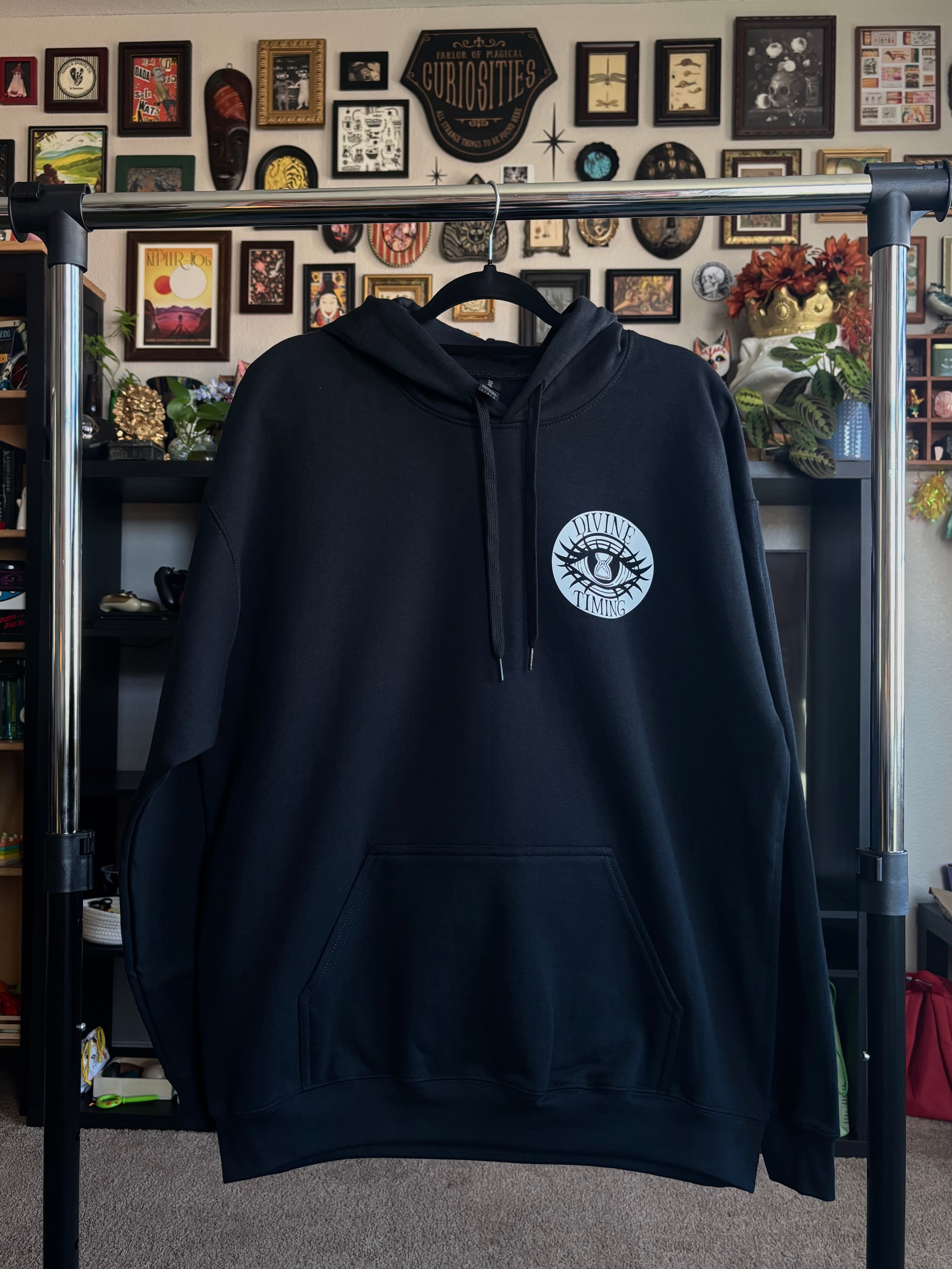 black hoodie hanging on a rack to show the front with the Divine Timing Hourglass Eye logo printed as a club patch