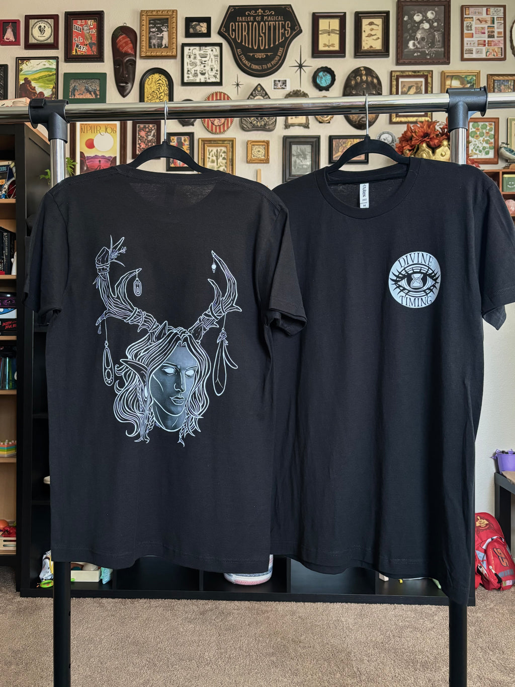 two black tees hanging on a rack to display the King of the Forest, a Celtic inspired fae lord, printed across the back and the Divine Timing Hourglass Eye printed as a club patch on the front