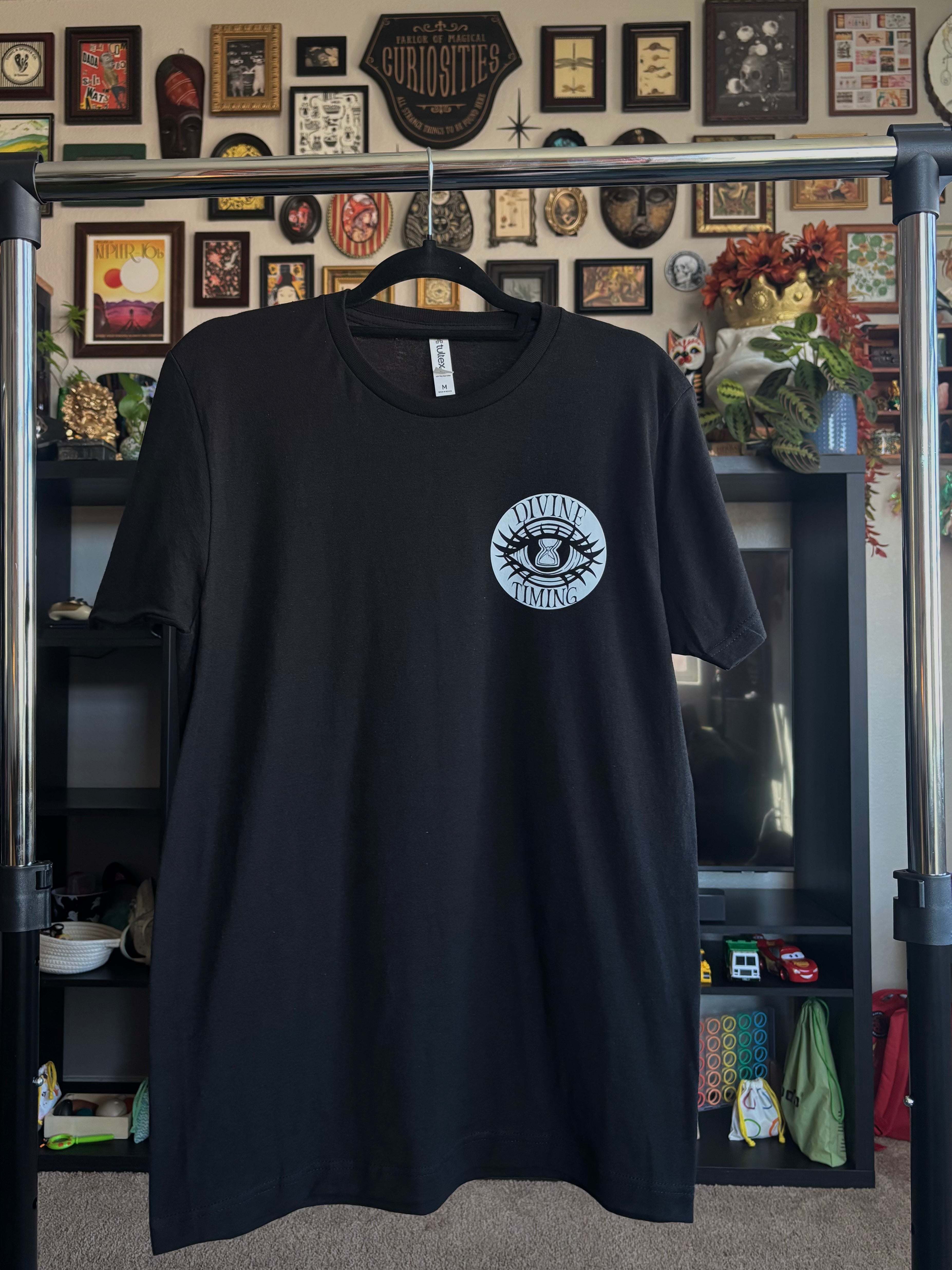 black tee hanging on a rack to show the front with the Divine Timing Hourglass Eye logo printed as a club patch