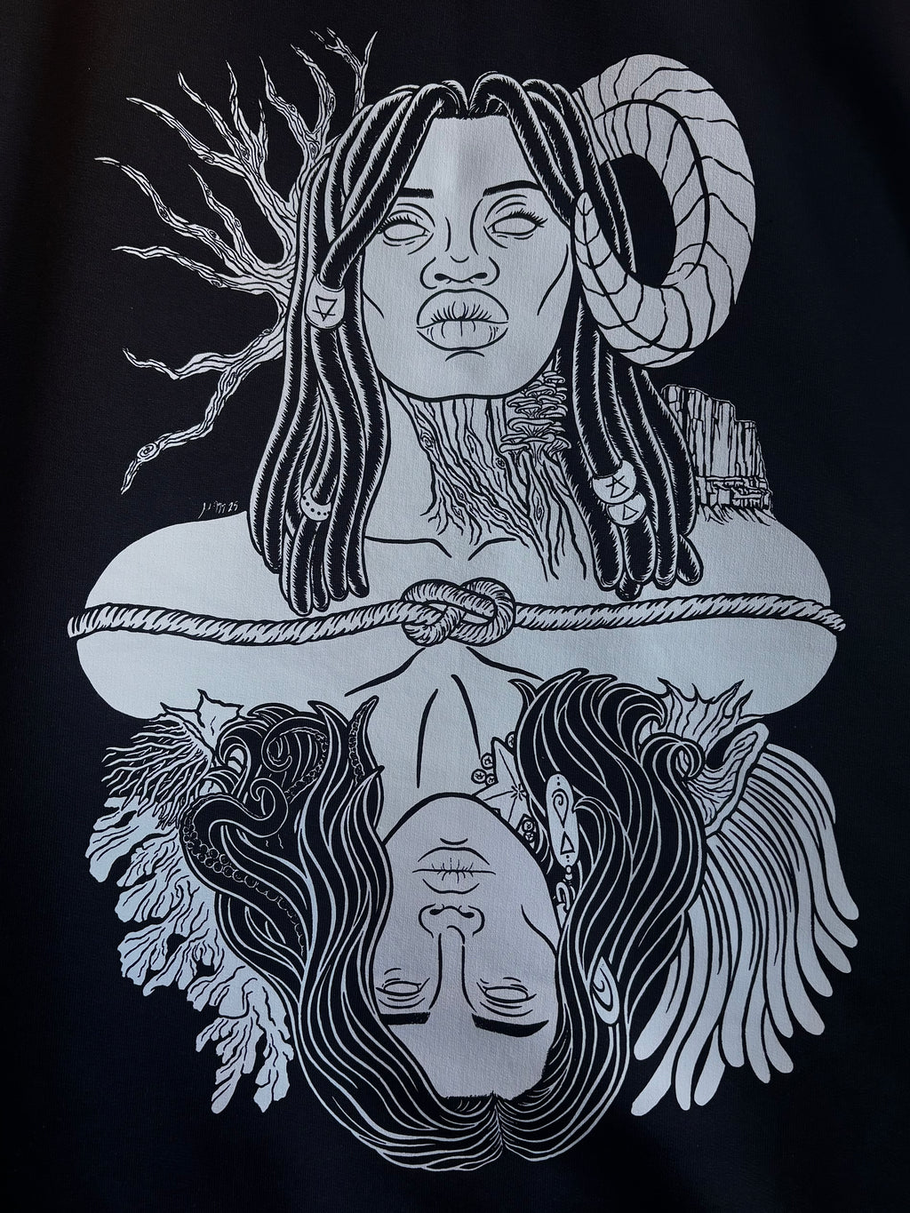 closeup look at the graphic design of Gaia, a powerful goddess adorned with beaded braids, a ram's horn, trees and mountains, who is connected to Oceanus, a god surrounded by coral and anemones with tentacles in his hair and a starfish and barnacles clinging to his neck, printed on the back of a black hoodie
