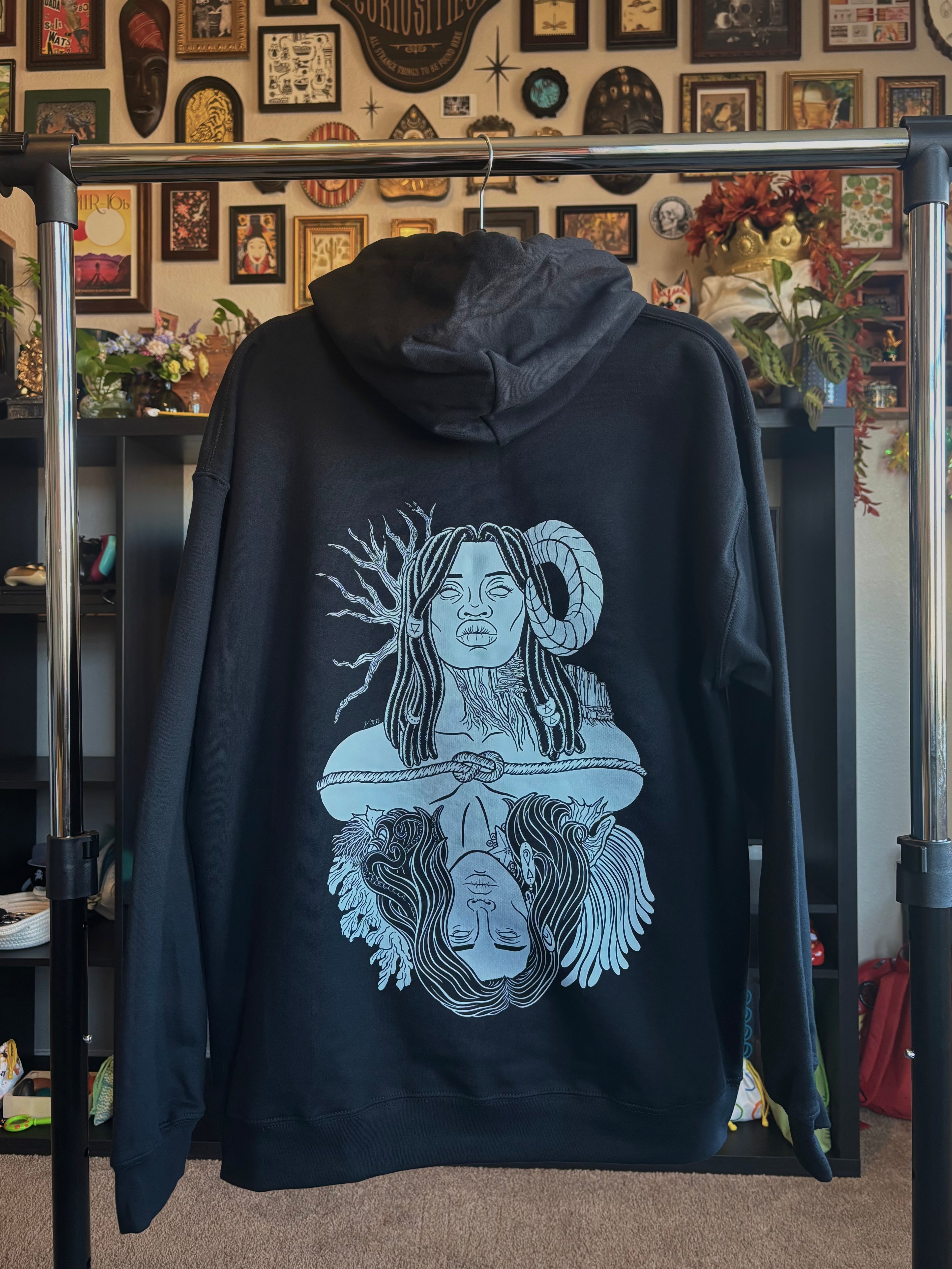 black hoodie hanging on a rack to show the full back print: a graphic design of Gaia, a powerful goddess adorned with beaded braids, a ram's horn, trees and mountains, who is connected to Oceanus, a god surrounded by coral and anemones with tentacles in his hair and a starfish and barnacles clinging to his neck