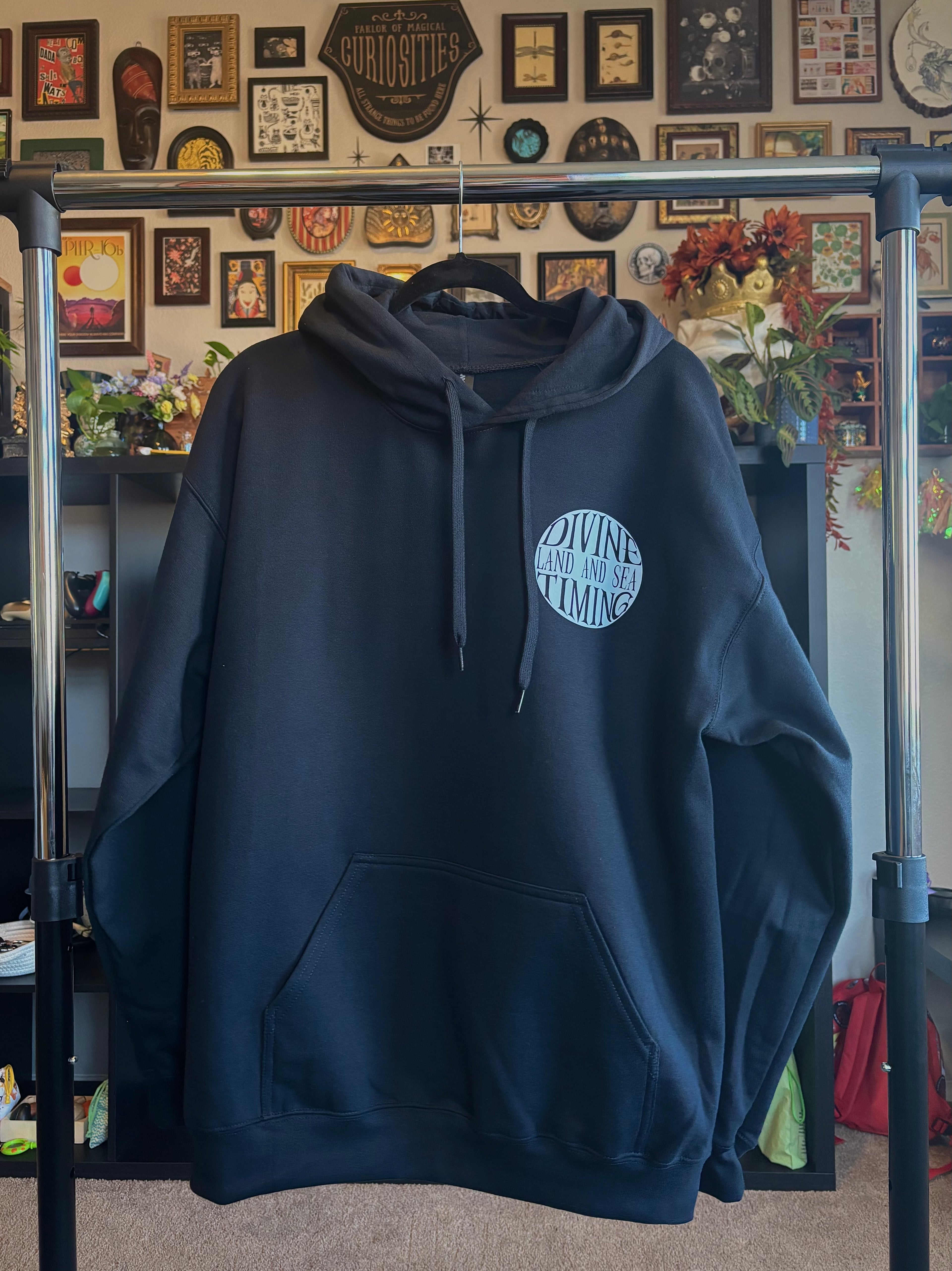 black hoodie hanging to show the front with the Divine Timing Land and Sea badge printed as a club patch