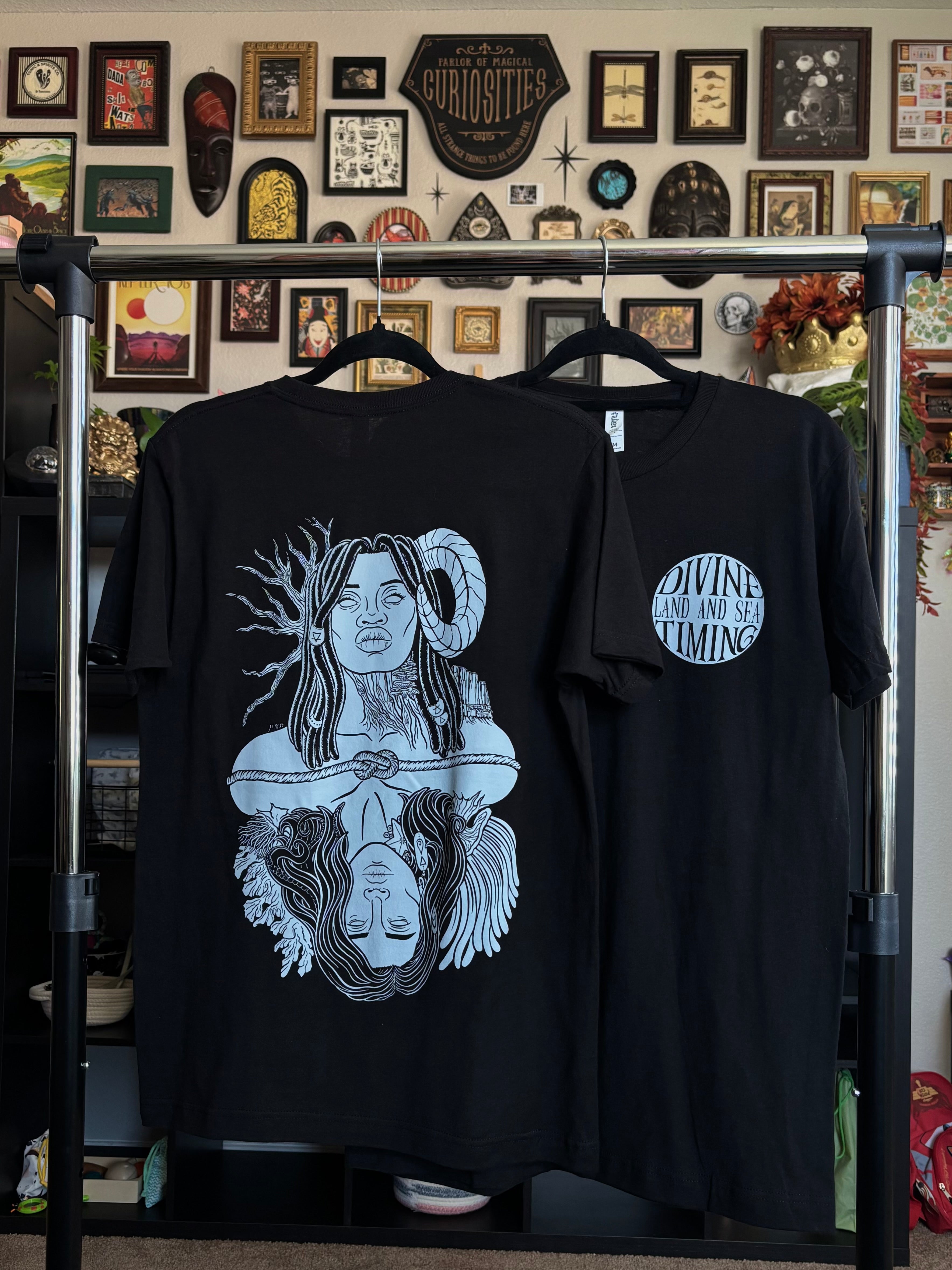 two black tees hanging on a rack to display the full back print of Gaia and Oceanus and the club patch on the front