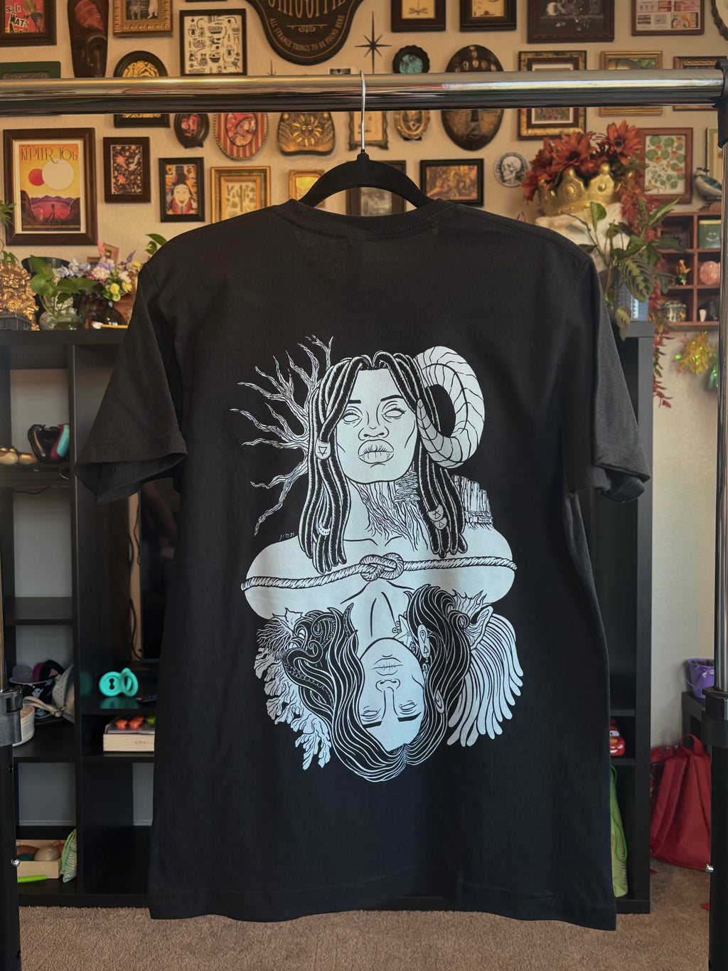black tee hanging on a rack to show the full back print: a graphic design of Gaia, a powerful goddess adorned with beaded braids, a ram's horn, trees and mountains, who is connected to Oceanus, a god surrounded by coral and anemones with tentacles in his hair and a starfish and barnacles clinging to his neck