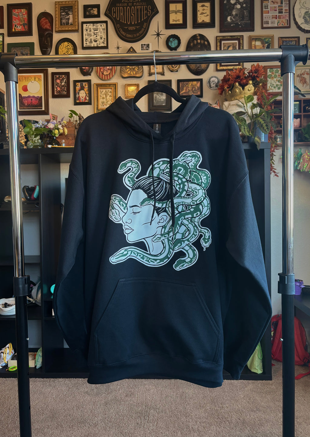 black hoodie with a graphic design of Medusa, shown in a side profile with several curling snakes, hanging on a rack