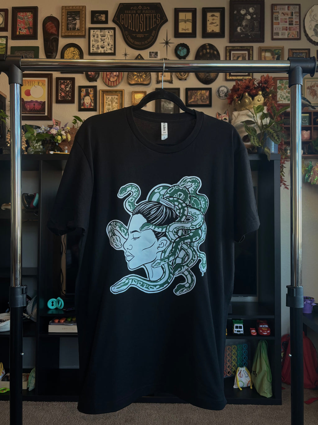 black tee with a graphic design of Medusa, shown in a side profile with several curling snakes, hanging on a rack