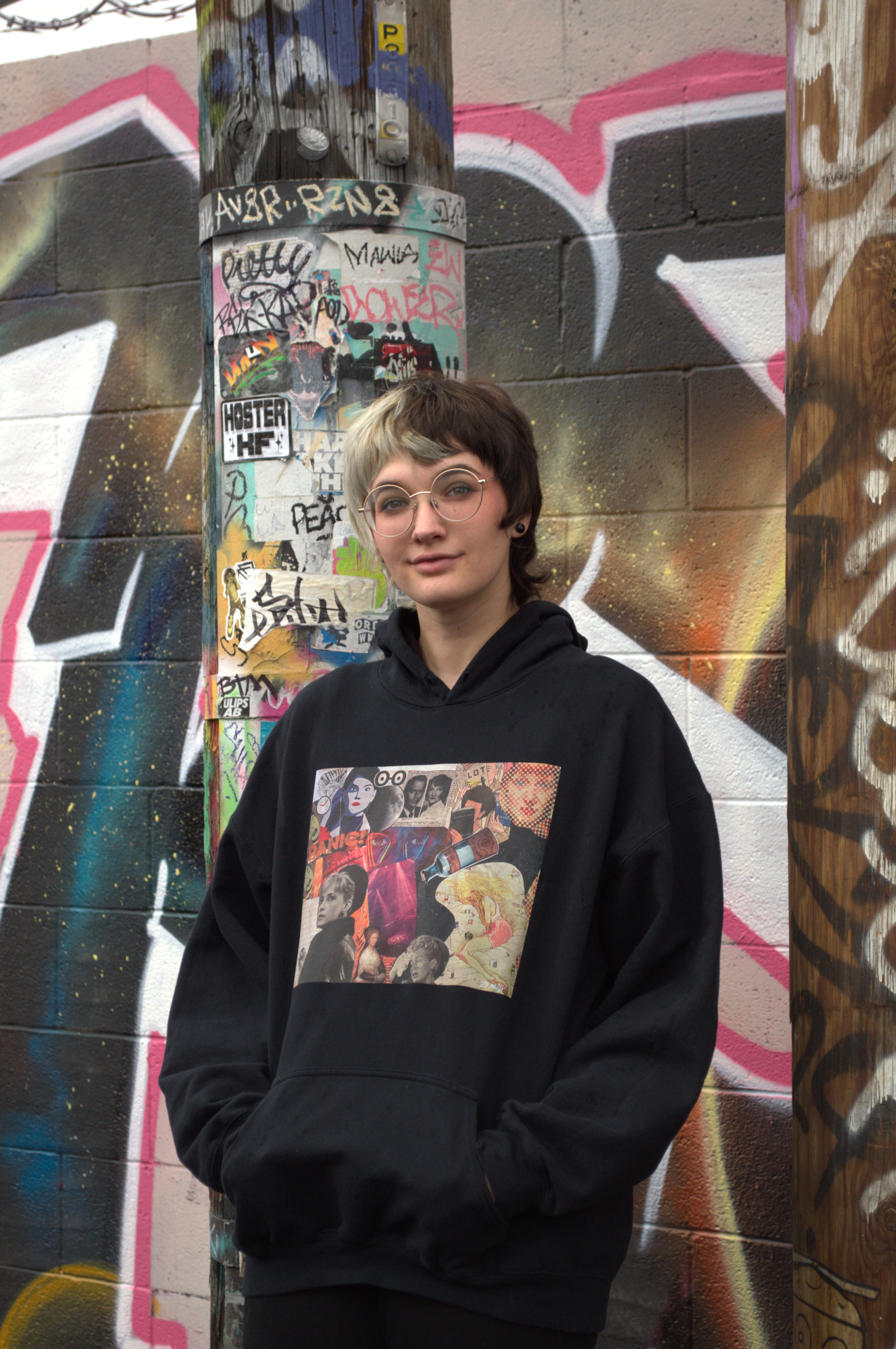 Person wearing a black hoodie with a colorful graphic design in front of a graffiti-covered wall.