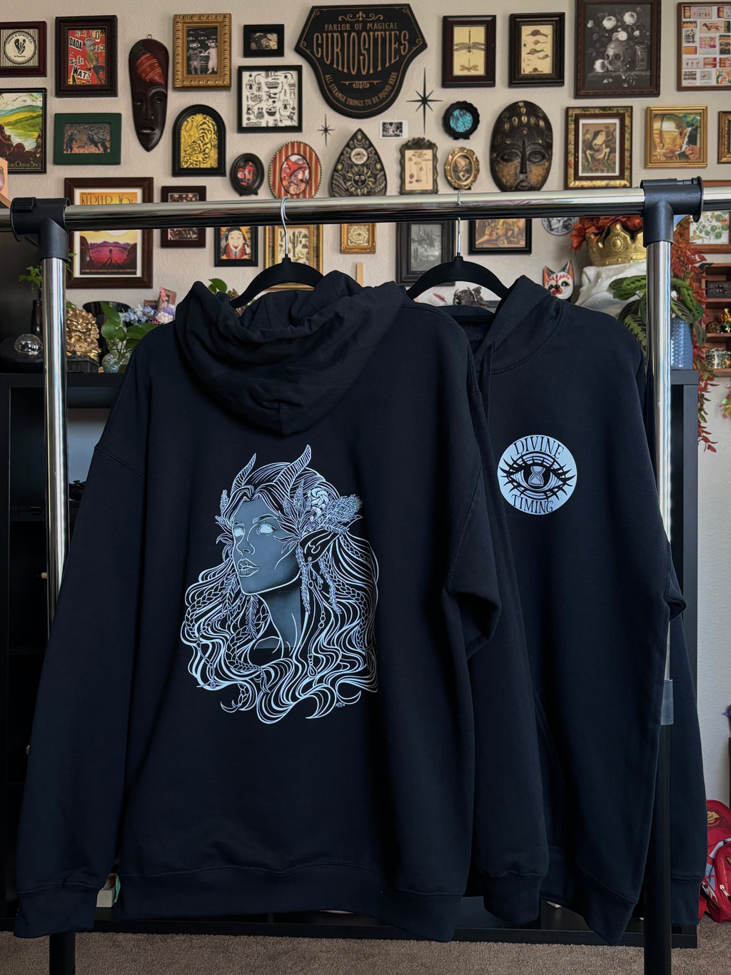 two black hoodies hanging on a rack to display the Queen of the Forest, a Celtic inspired fae goddess, printed across the back and the Divine Timing Hourglass Eye printed as a club patch on the front