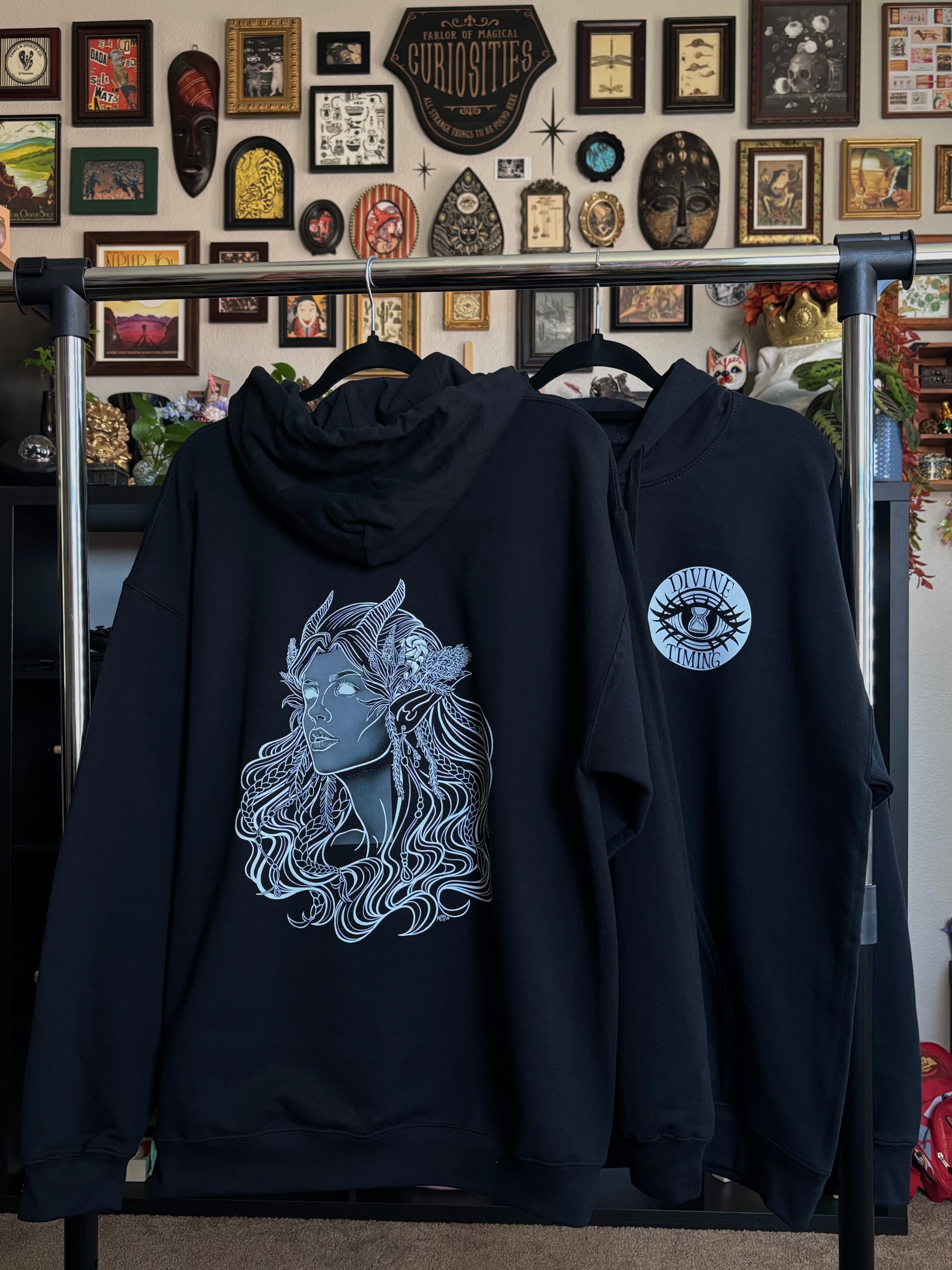 two black hoodies hanging on a rack to display the Queen of the Forest, a Celtic inspired fae goddess, printed across the back and the Divine Timing Hourglass Eye printed as a club patch on the front