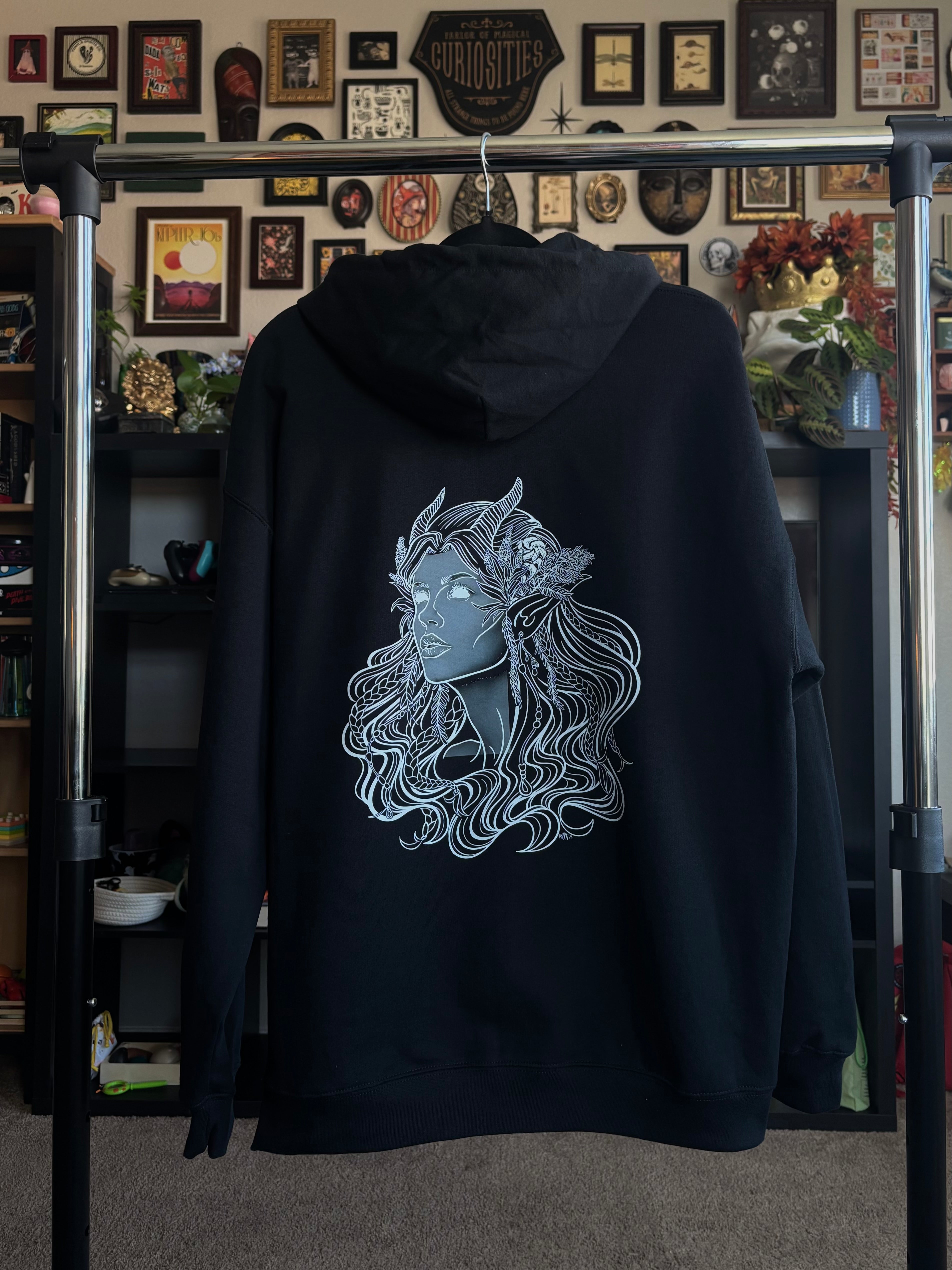 black hoodie hanging on a rack to display the Queen of the Forest, a Celtic inspired fae goddess, printed across the back