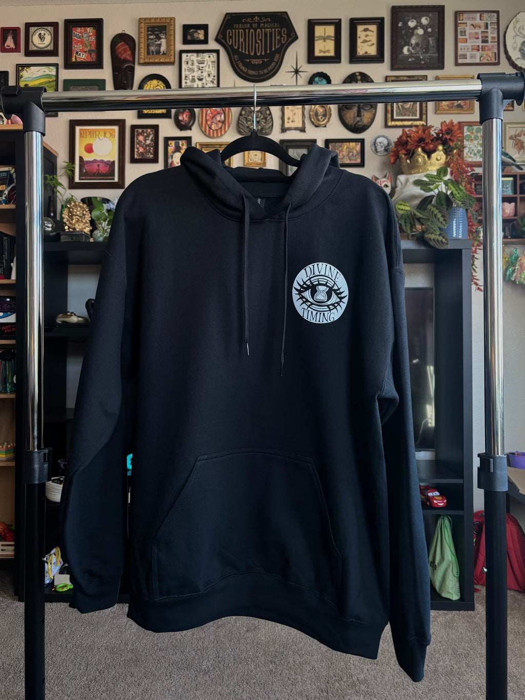 black hoodie hanging on a rack to show the front with the Divine Timing Hourglass Eye logo printed as a club patch