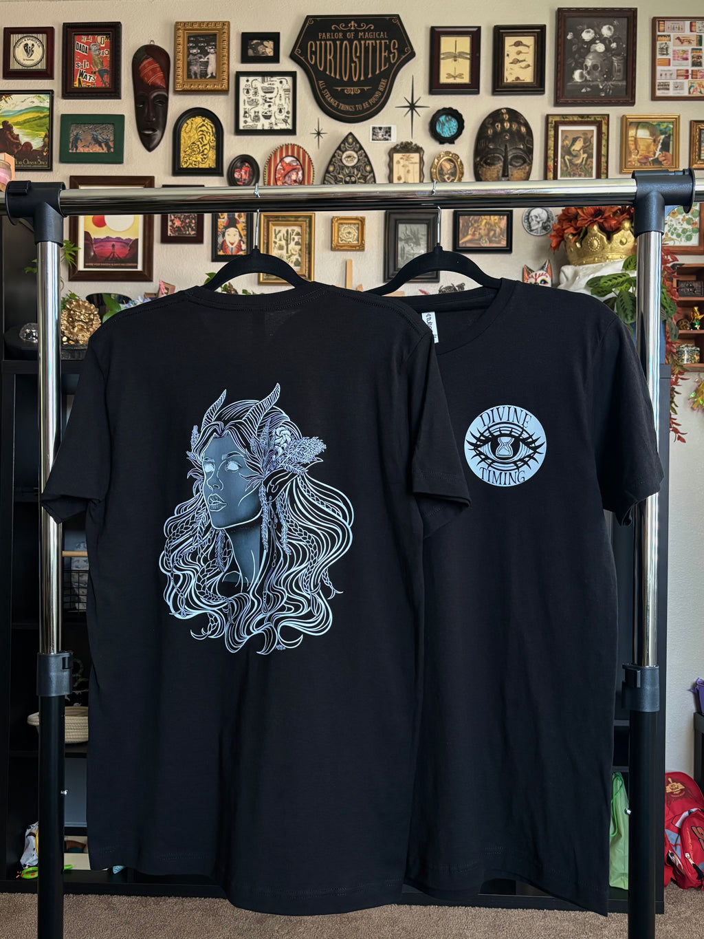 two black tees hanging on a rack to display the Queen of the Forest, a Celtic inspired fae goddess, printed across the back and the Divine Timing Hourglass Eye printed as a club patch on the front