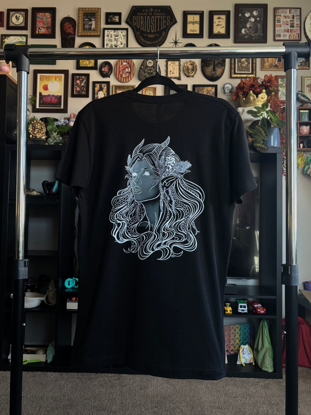 black tee hanging on a rack to display the Queen of the Forest, a Celtic inspired fae goddess, printed across the back