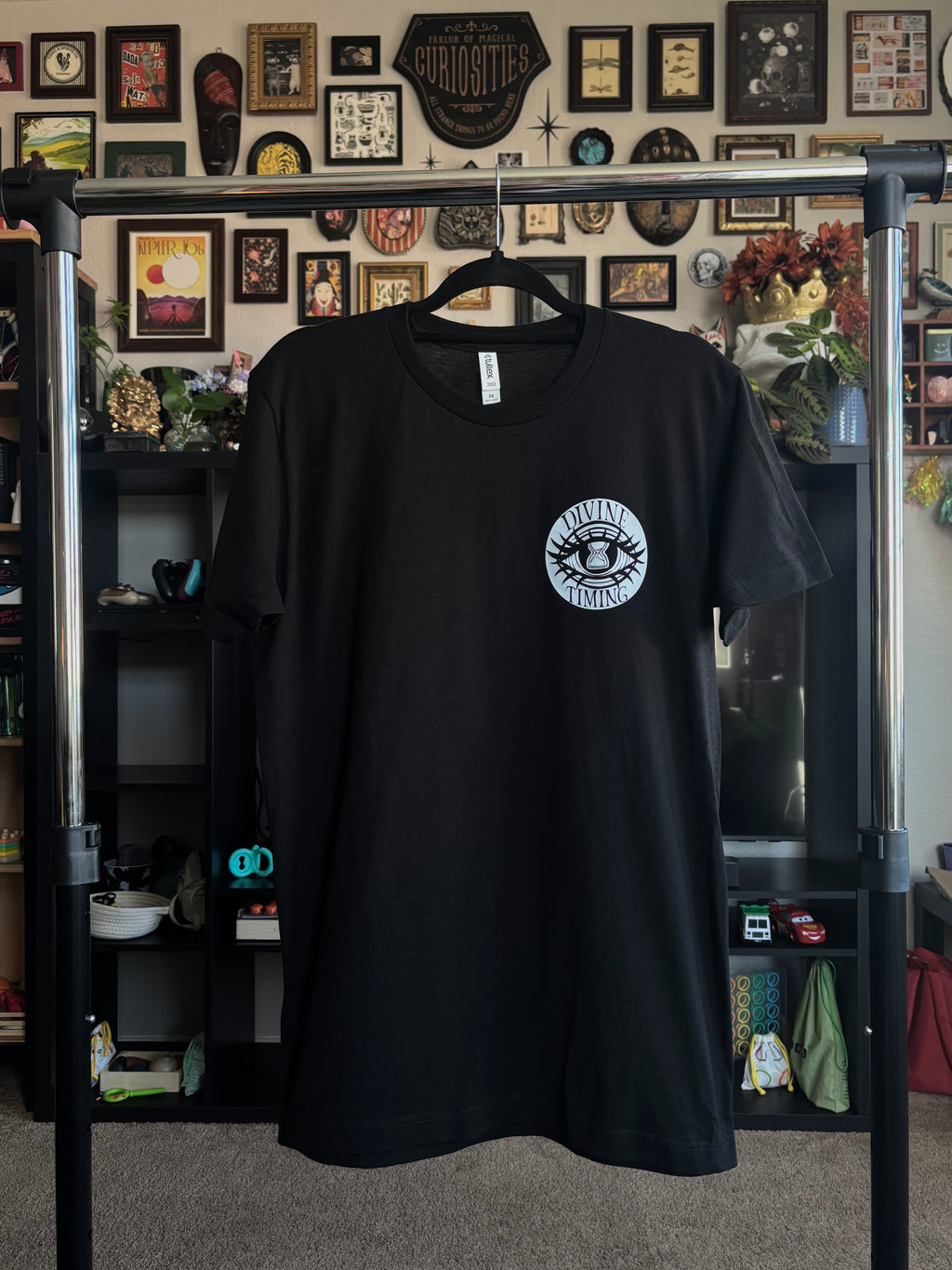 black tee hanging on a rack to show the front with the Divine Timing Hourglass Eye logo printed as a club patch